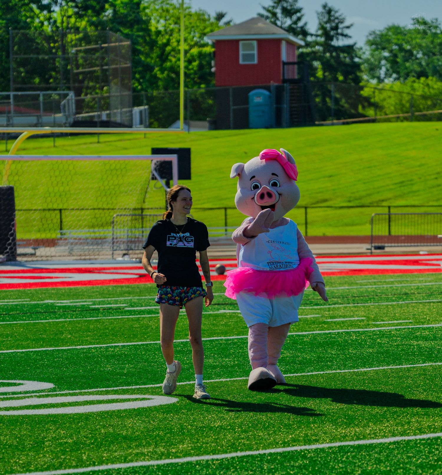A girl walks alongside a pink pig mascot in a tutu on a green turf football field on a sunny day.