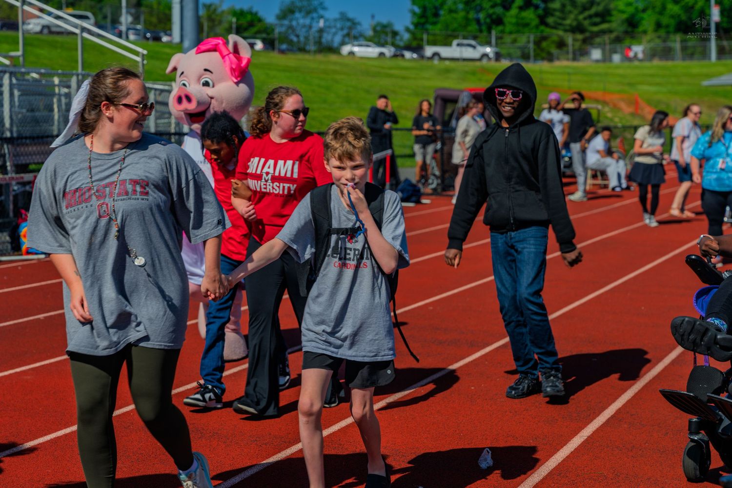 People walking on a red track at a school event with a pink pig mascot costume visible in the background.