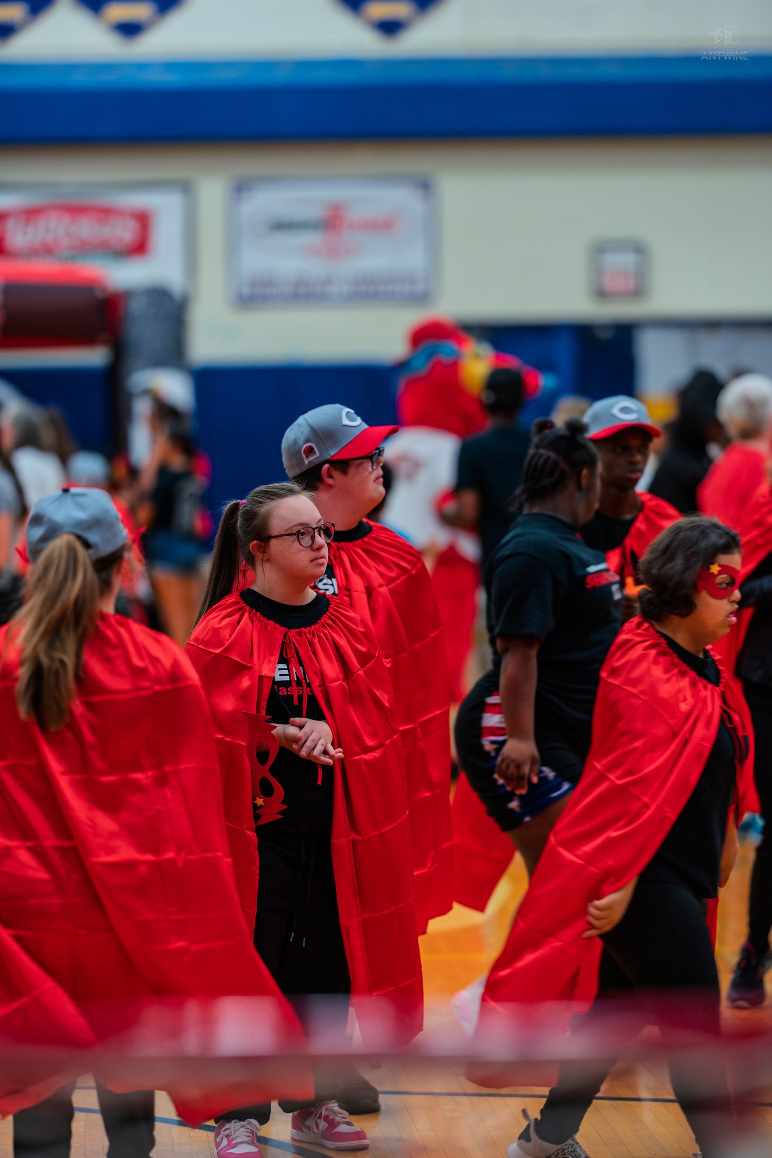 Group of Special Olympics athletes wearing red superhero capes walk together inside a gymnasium during an event.