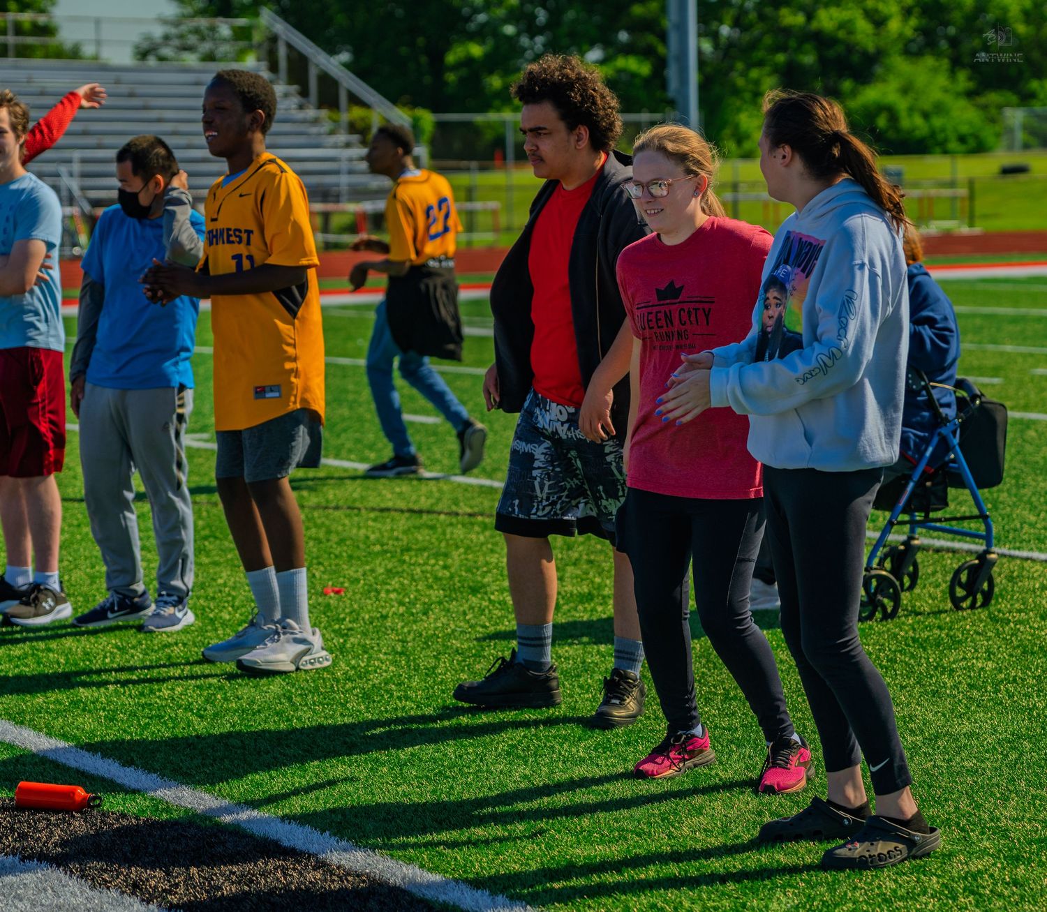 Group of people participating in outdoor track and field activities on a green turf sports field on a sunny day.