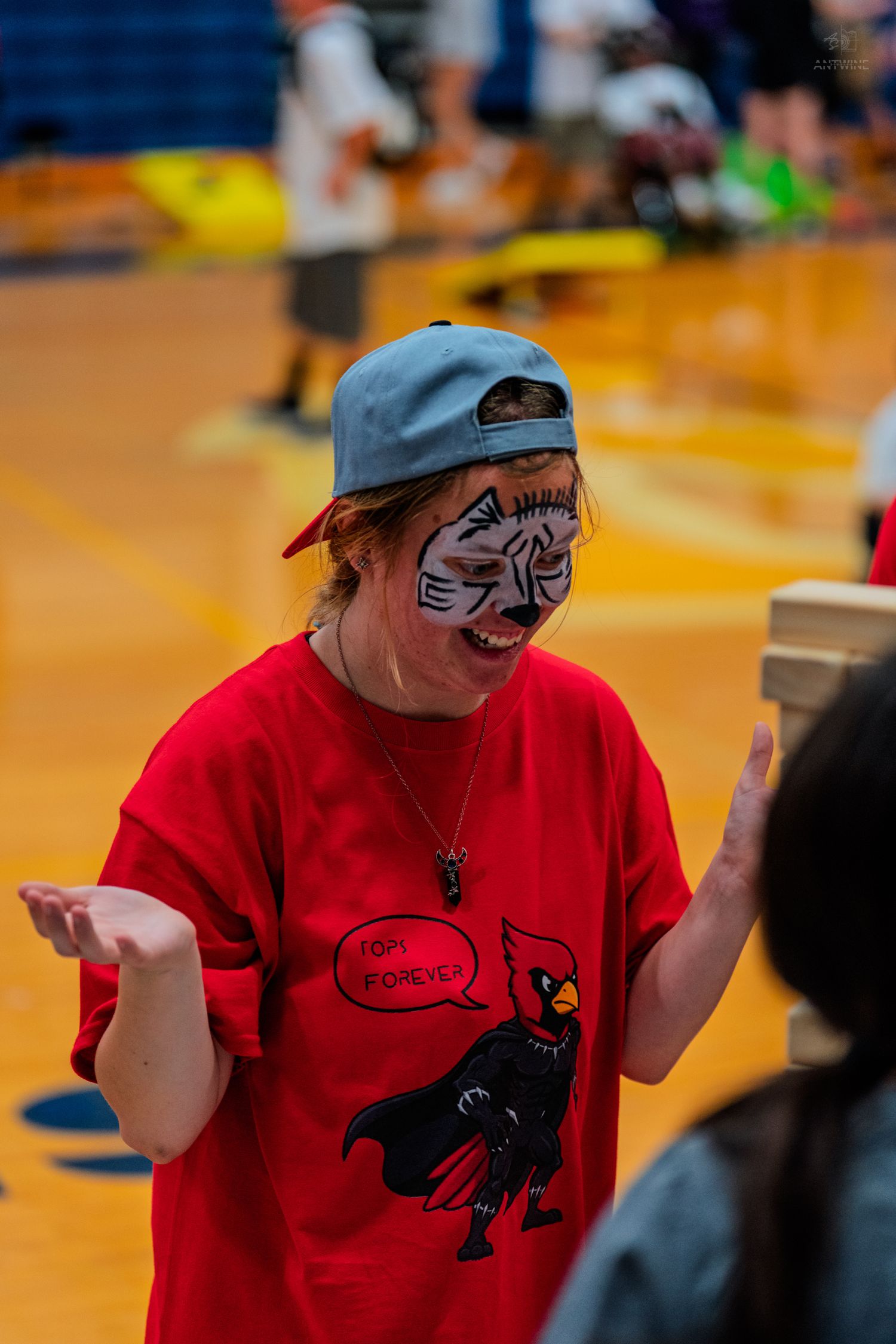 Child with cat face paint and backward cap wearing red t-shirt gestures expressively at indoor gym event.