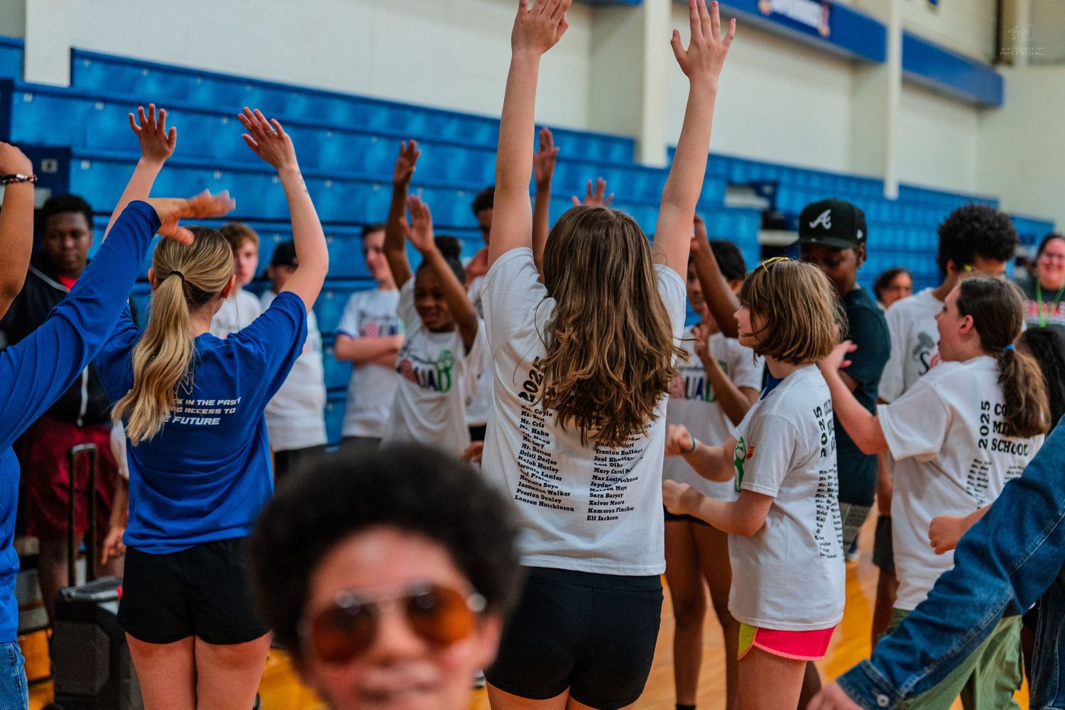 Group of teens in white t-shirts raising hands at an indoor gym event or school rally.