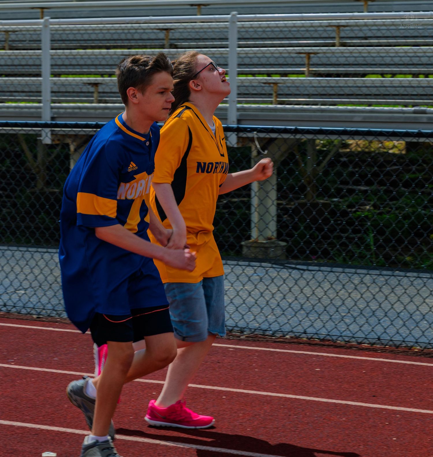 Two youth athletes in blue and gold jerseys run together on a red track during a school sports event.