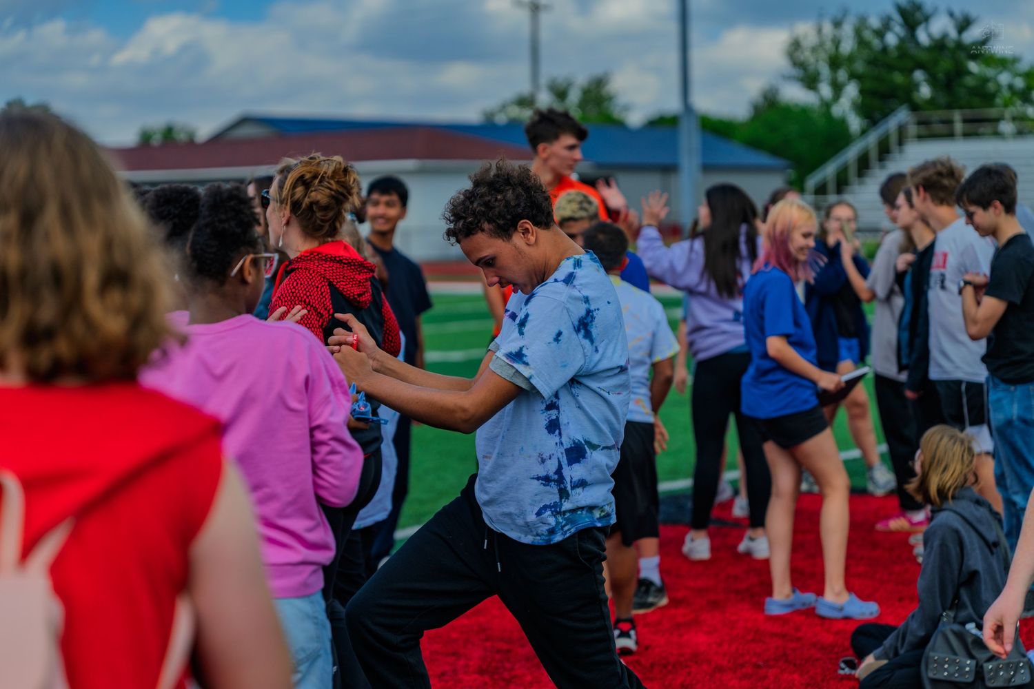 Students dance and socialize on a red turf field during an outdoor school event on a cloudy day.