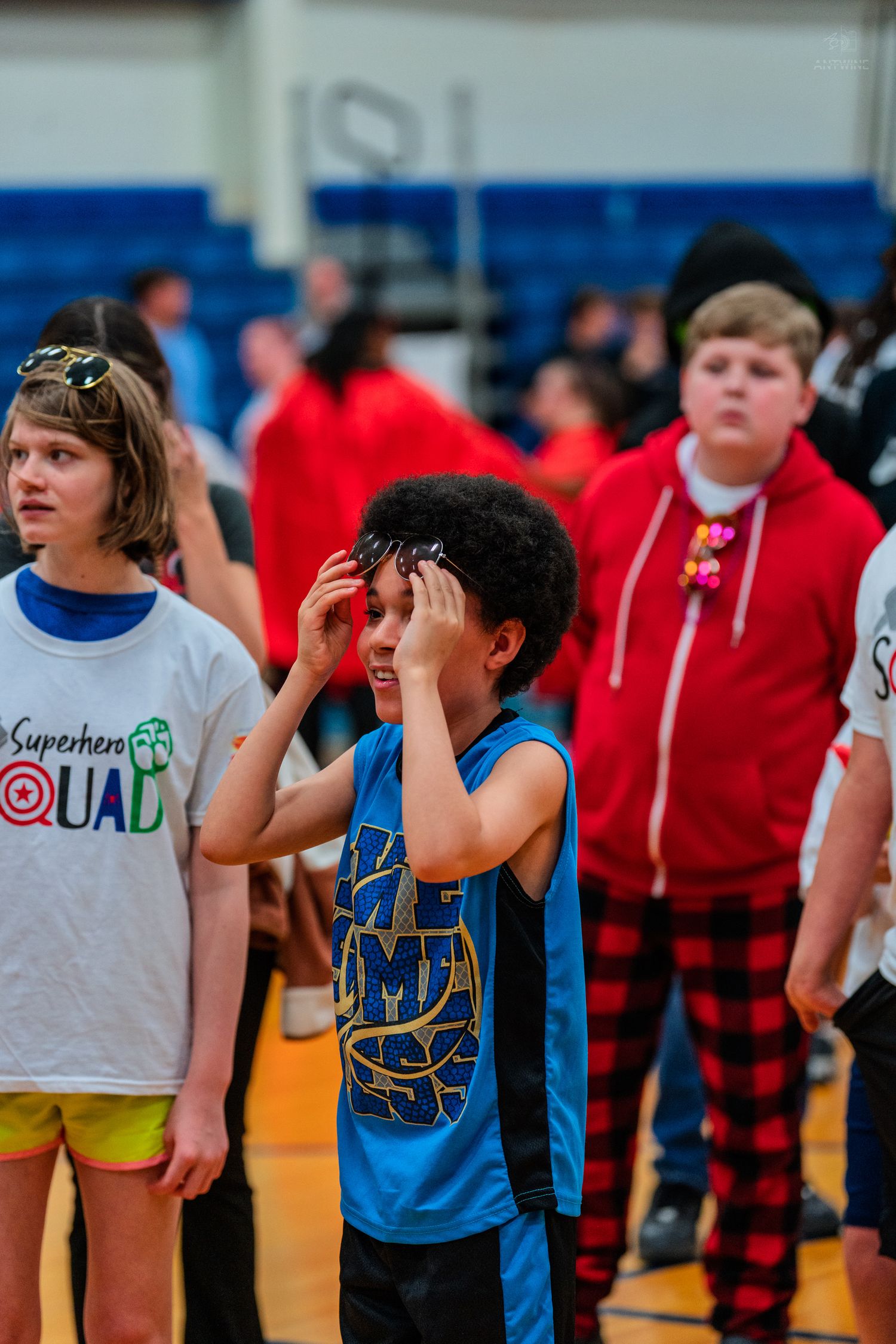 Children standing in a school gymnasium during an event, with one boy in a blue jersey shielding his eyes.