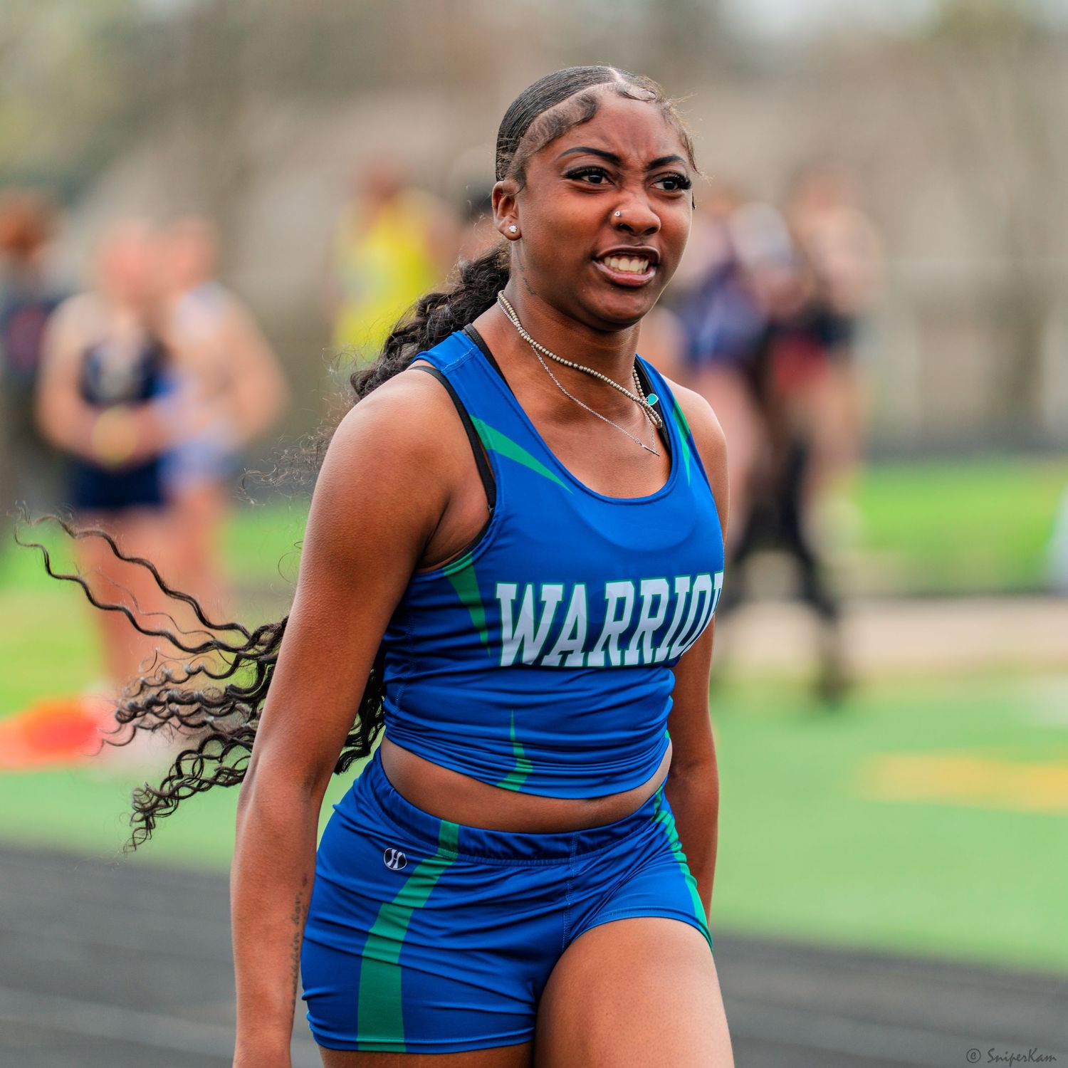 Female Warriors sprinter in blue uniform celebrates after finishing race at outdoor track meet.