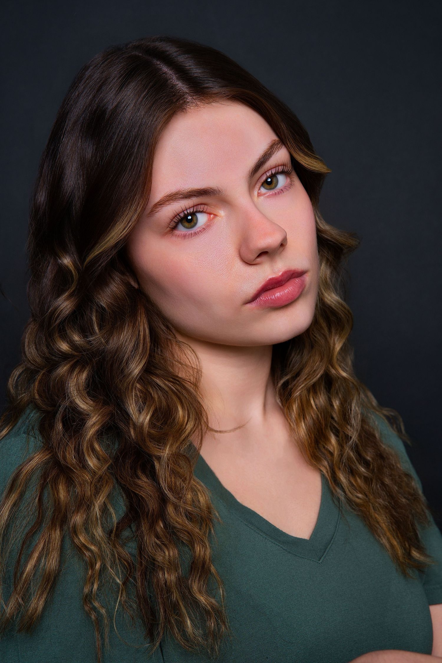 Young woman with long curly brown hair wearing a green v-neck shirt poses for a professional headshot against a dark background.