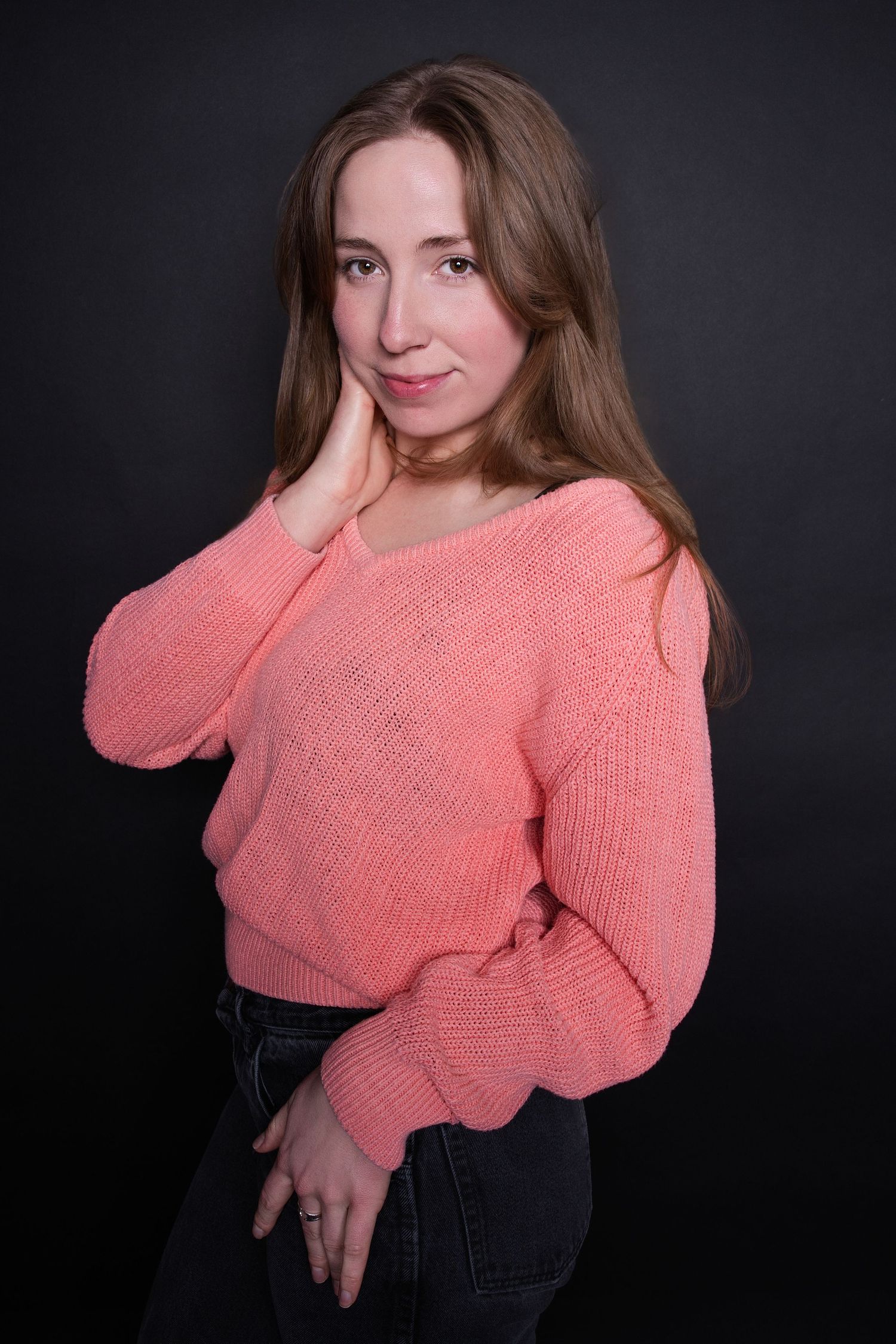 Woman in a pink knit sweater posing with hand to cheek against a dark studio background.