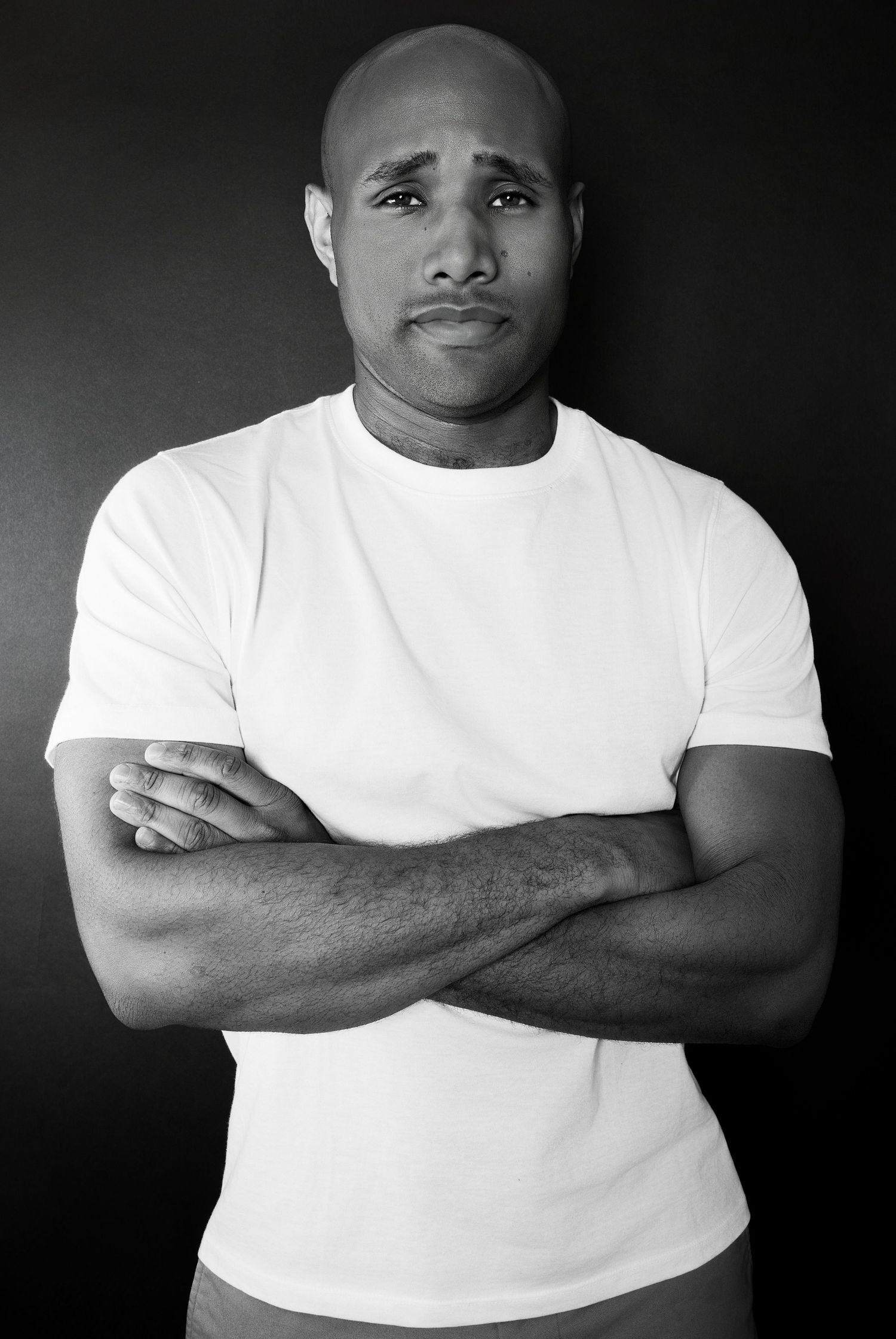 Bald Black man in white t-shirt with sad expression and arms crossed in black and white photo.
