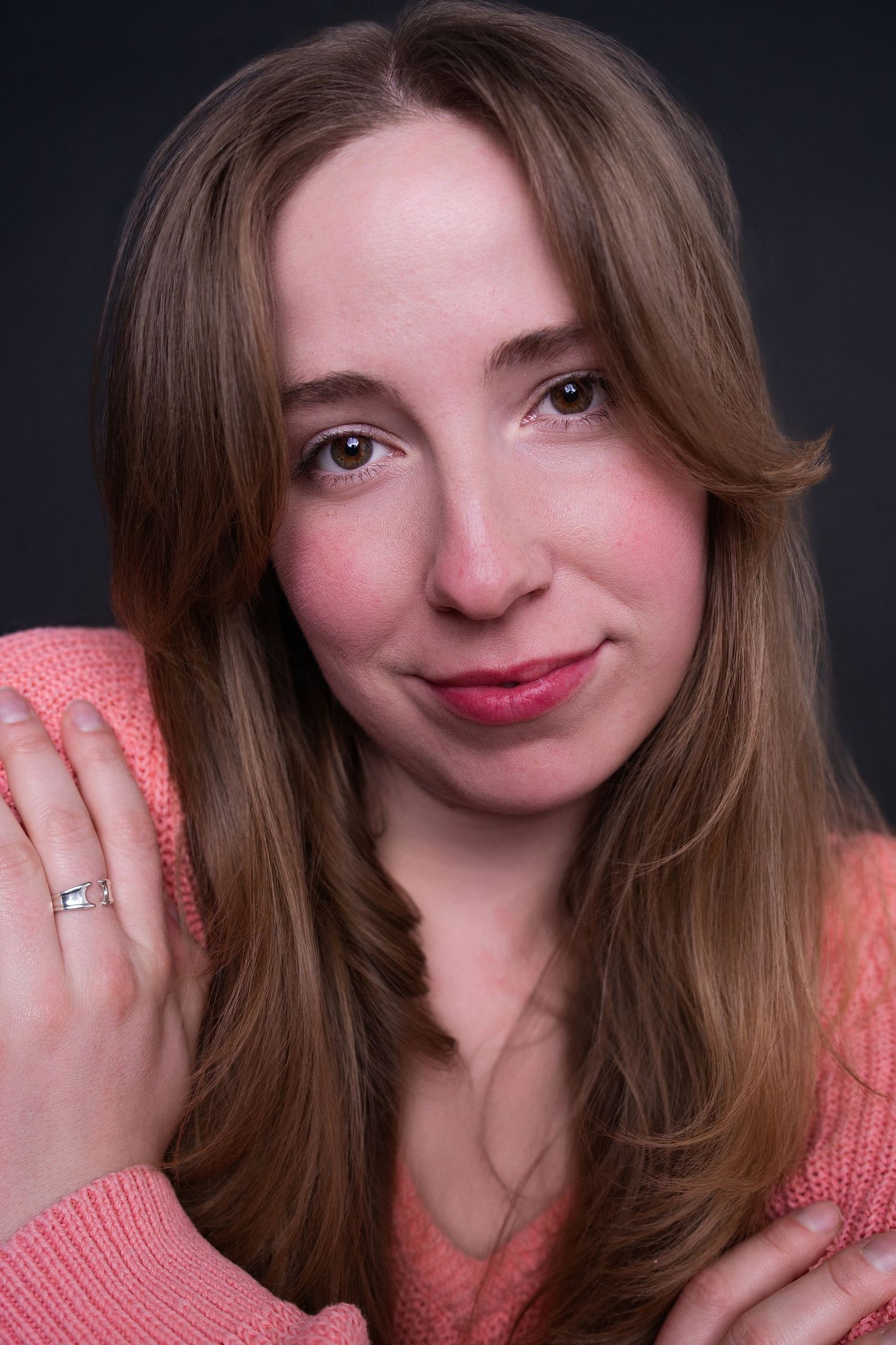Young woman in pink knit sweater with ring, arms crossed, smiling gently against dark gray background.