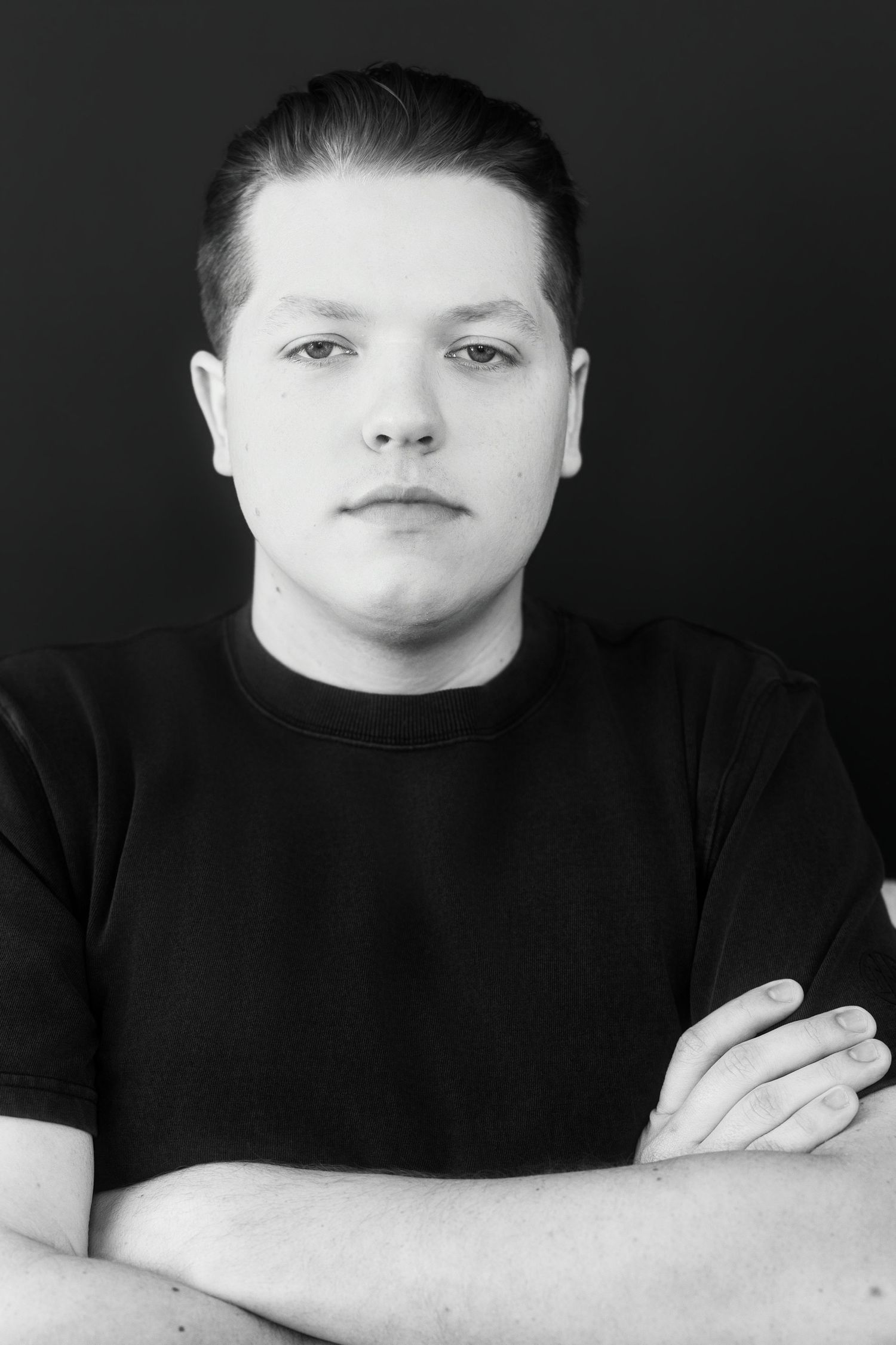 Black and white portrait of young man in black t-shirt with arms crossed against dark background.