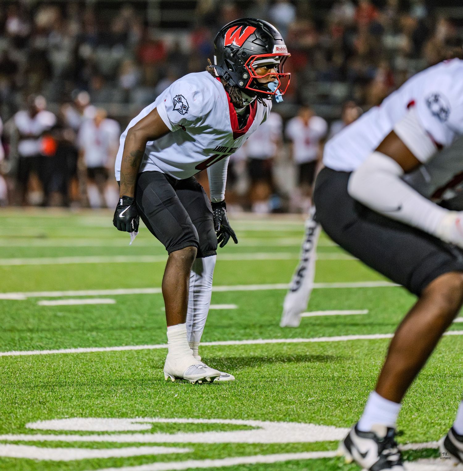 High school football player in black and red uniform assumes defensive stance on the field before a play.
