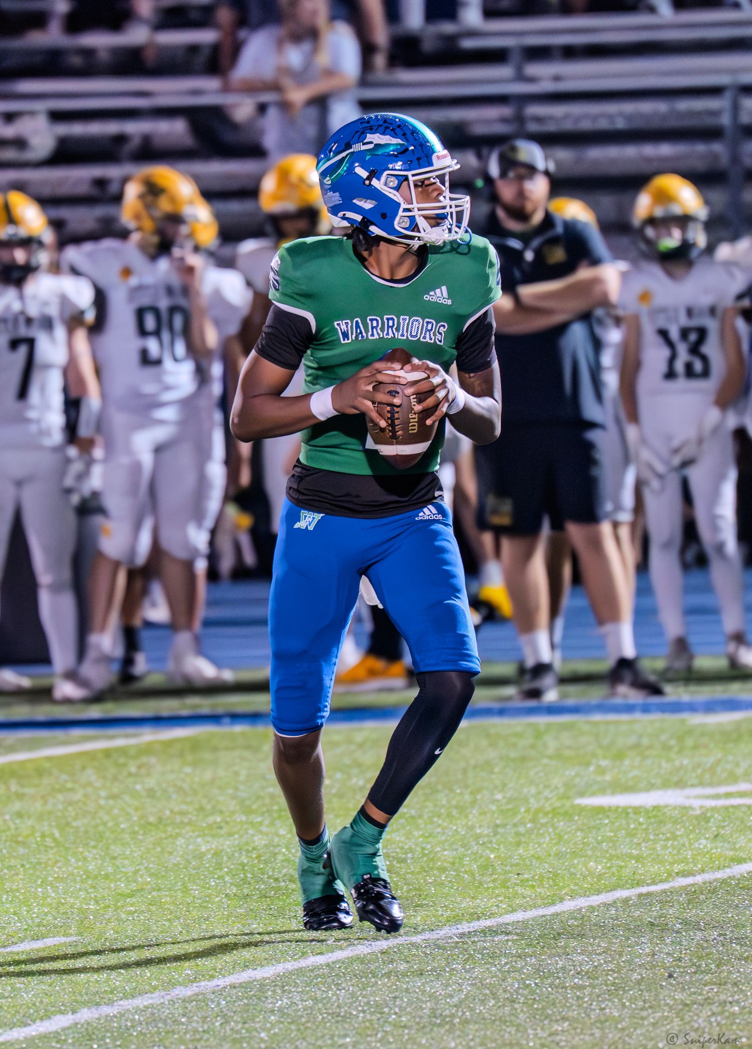 Warriors quarterback in green and blue uniform drops back to pass during a high school football game.