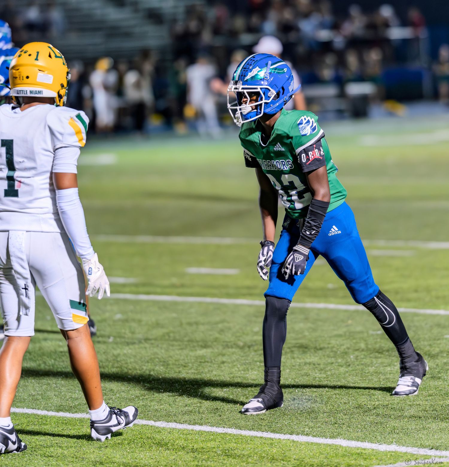 A football player in green and blue uniform stands in ready position facing an opponent in gold and white uniform.