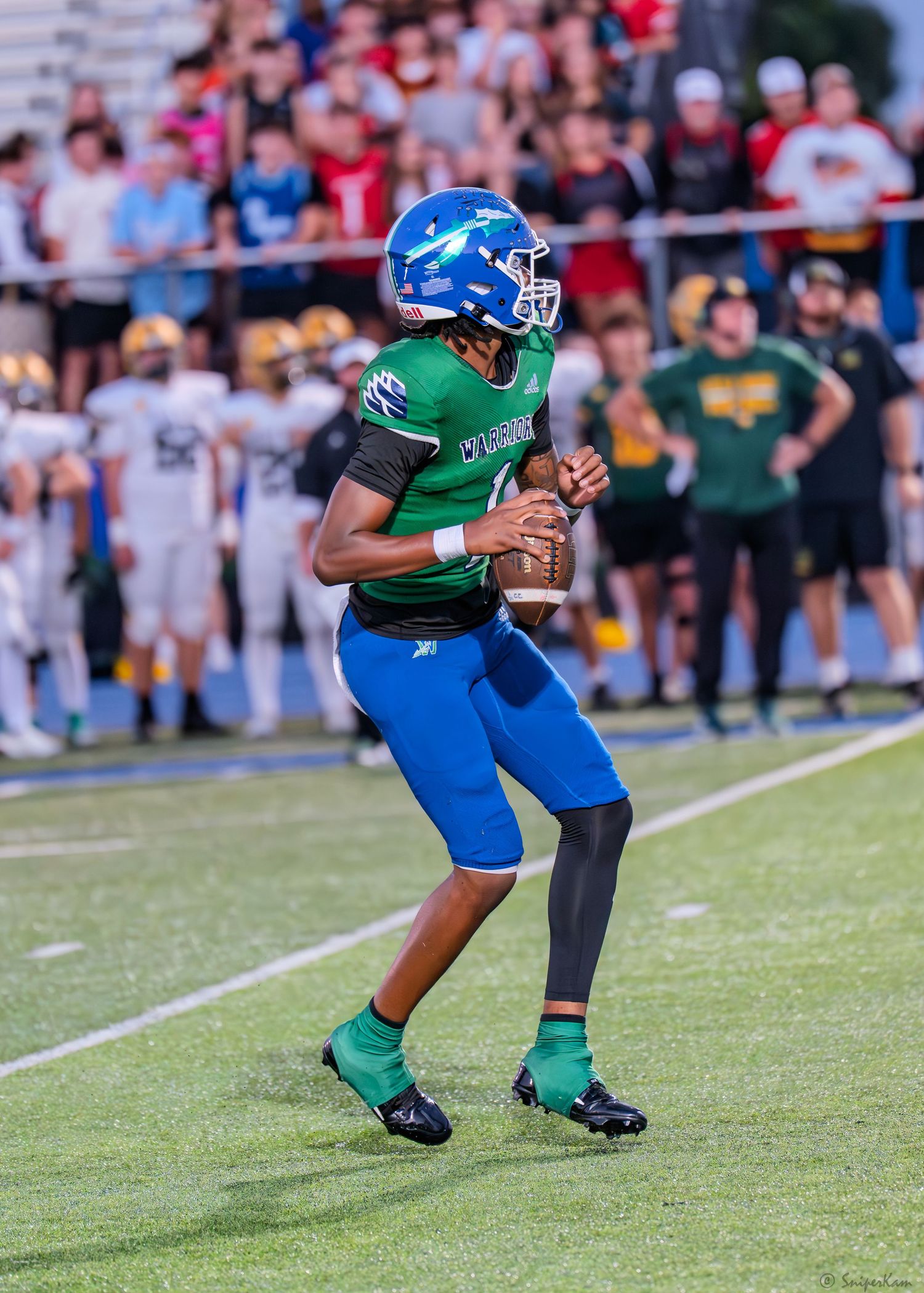 High school quarterback in blue and green uniform drops back to pass during a night football game.