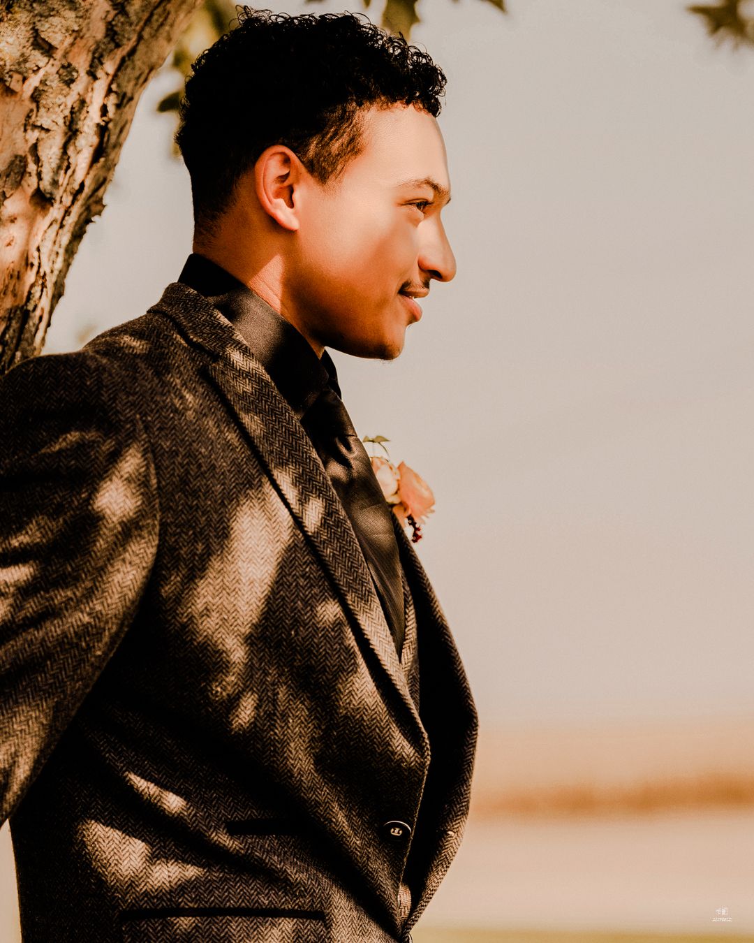 Groom in a dark suit with boutonniere leaning against a tree in a serene outdoor wedding portrait.