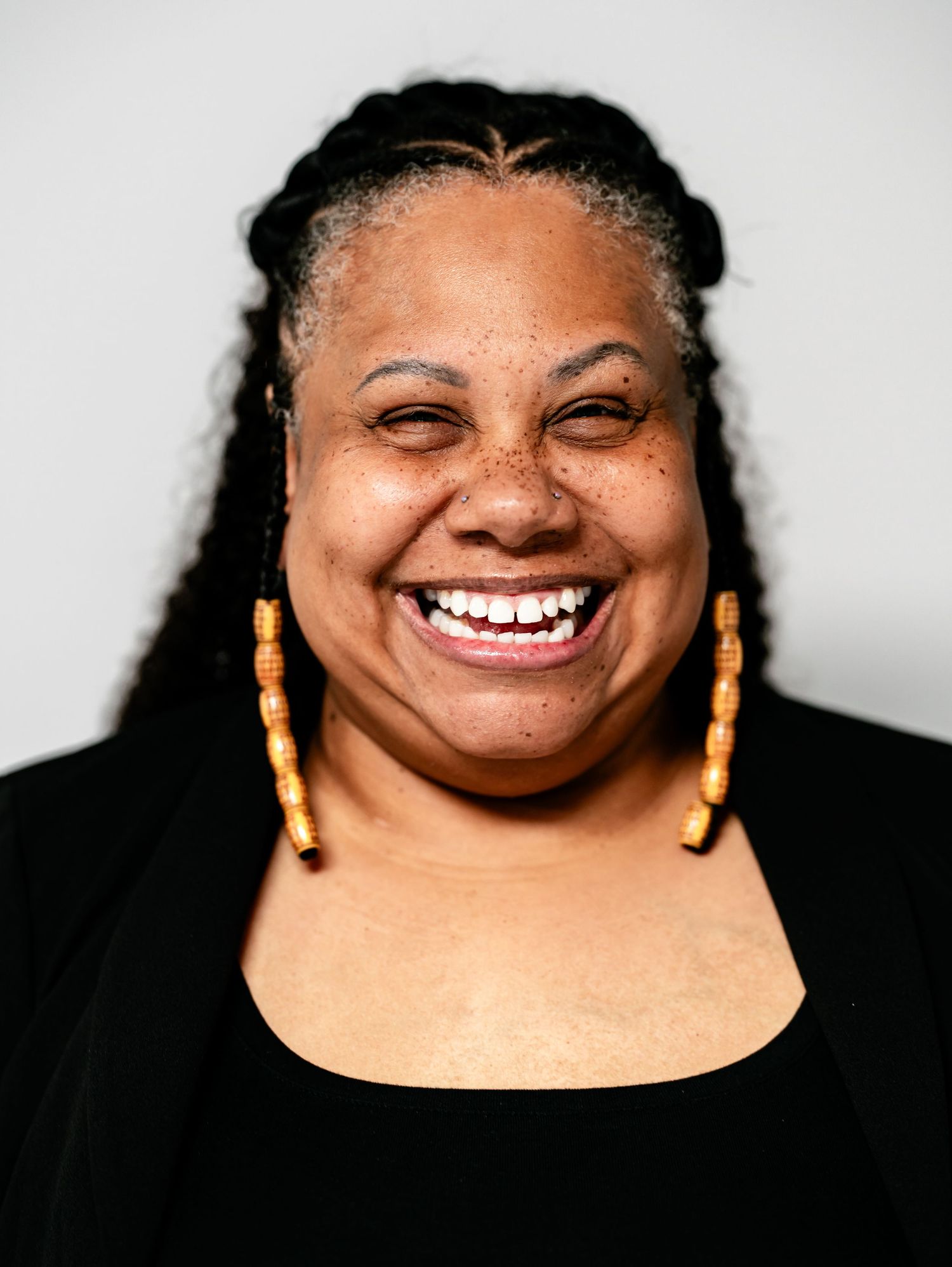 A Black woman with long braids and gold beaded earrings laughs broadly in a vibrant color portrait photo.