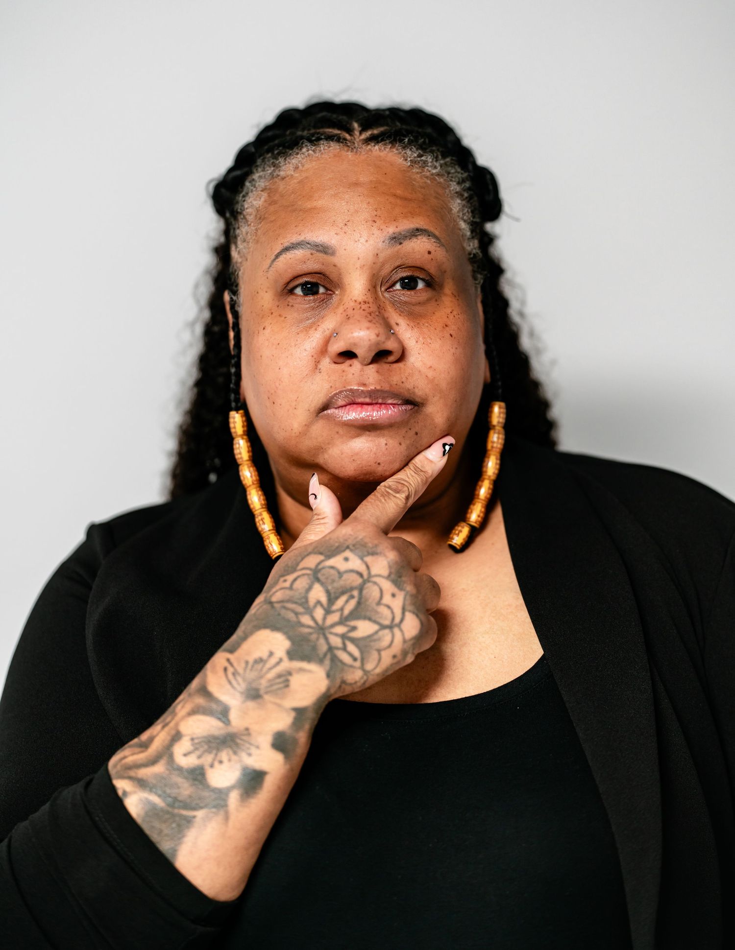 Woman with salt-and-pepper braids and floral hand tattoo poses in black outfit with gold earrings.