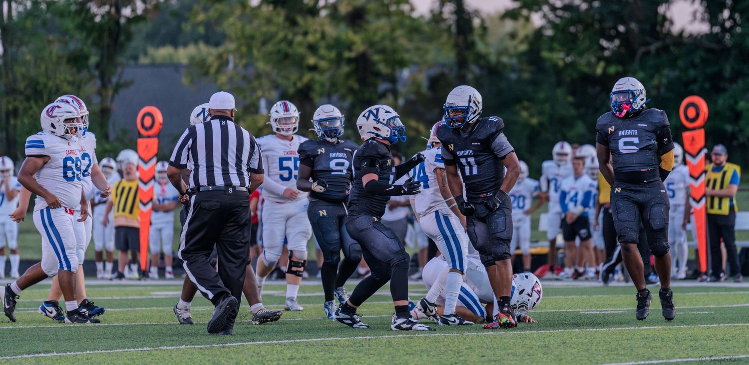 High school football players in black uniforms compete during a night game as a referee observes the play.