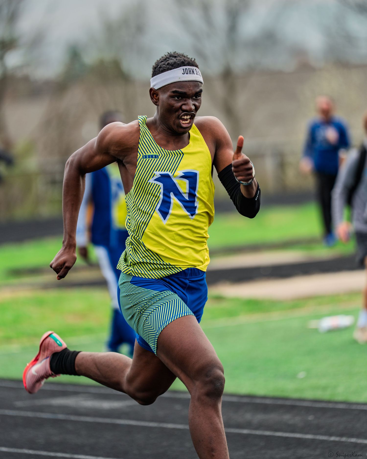 Male sprinter in yellow N jersey gives thumbs up while racing at outdoor track meet.