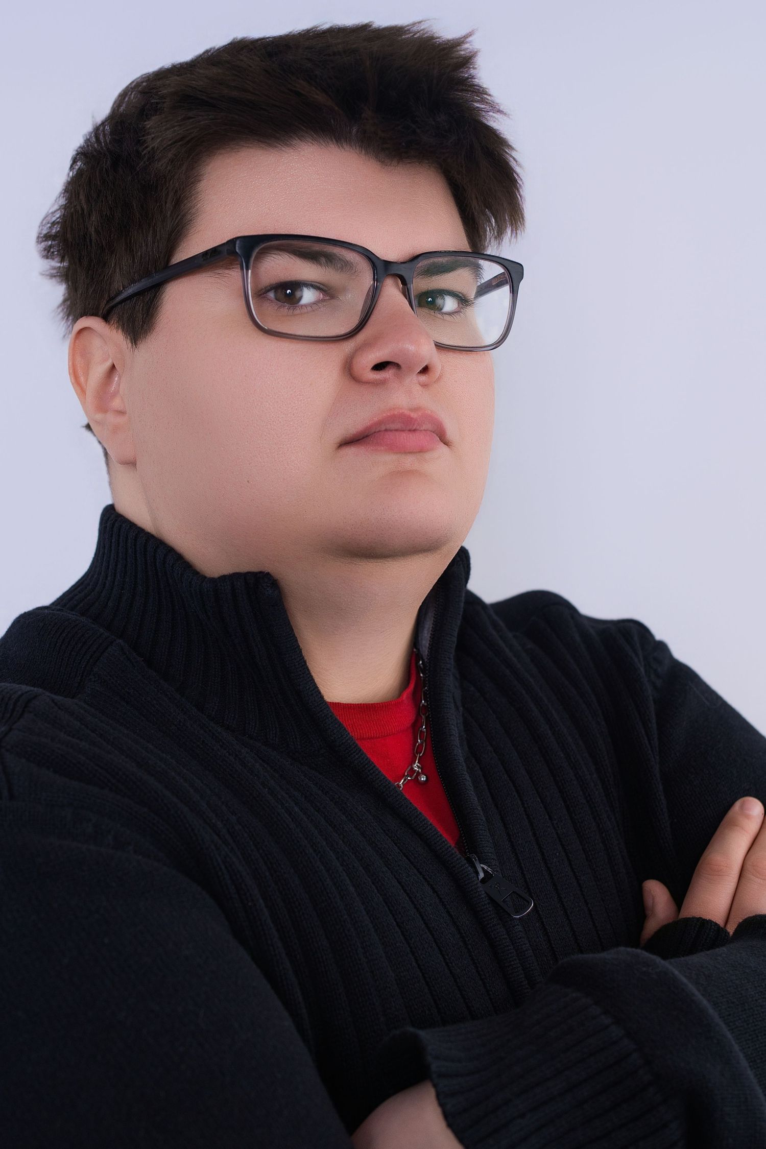 Young person in black zip sweater with arms crossed gazes upward in studio portrait.