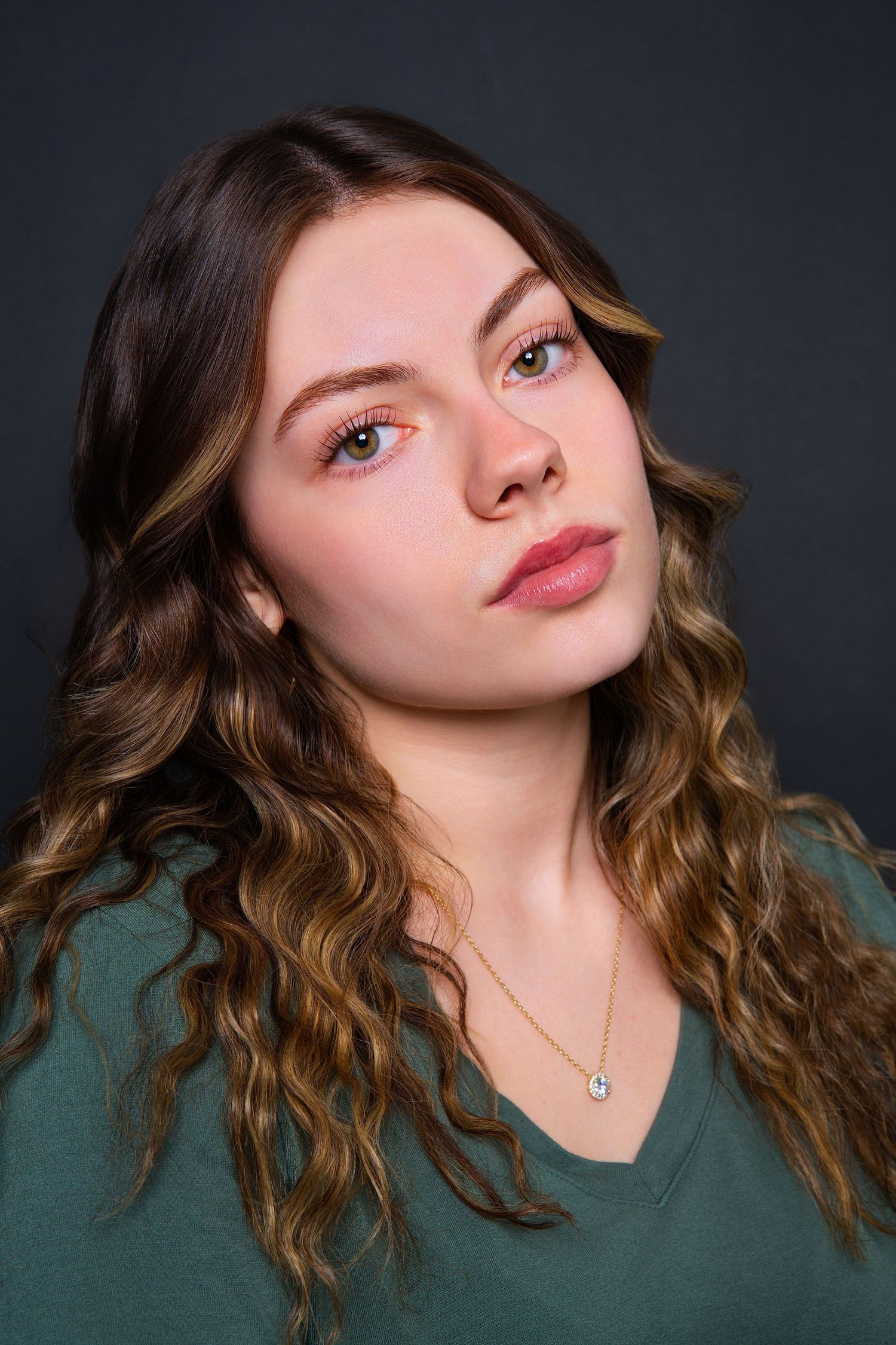 Young woman with long curly brown highlighted hair gazing upward in green v-neck top.