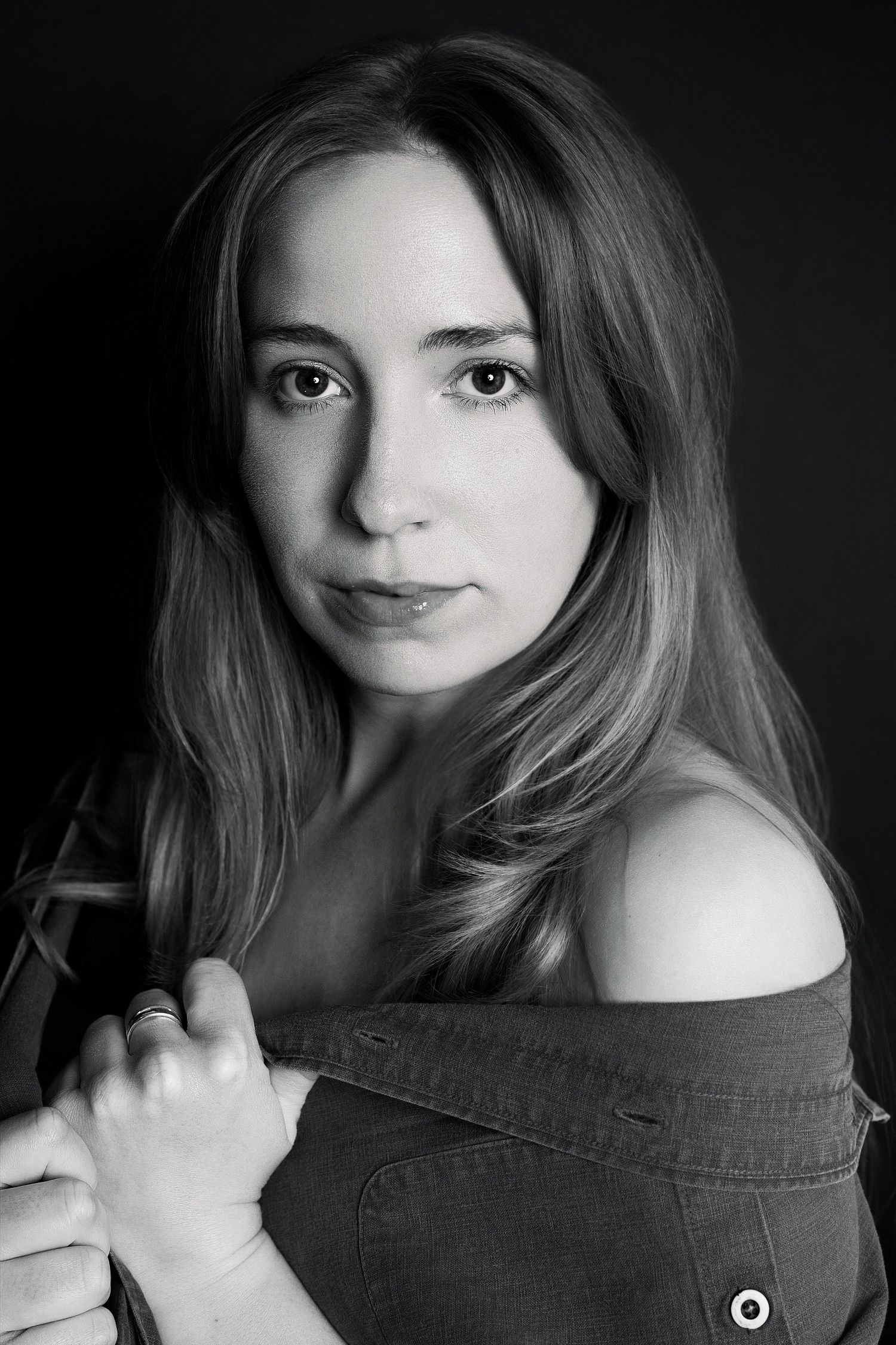Black and white portrait of young woman with long hair holding jacket off-shoulder against dark background.