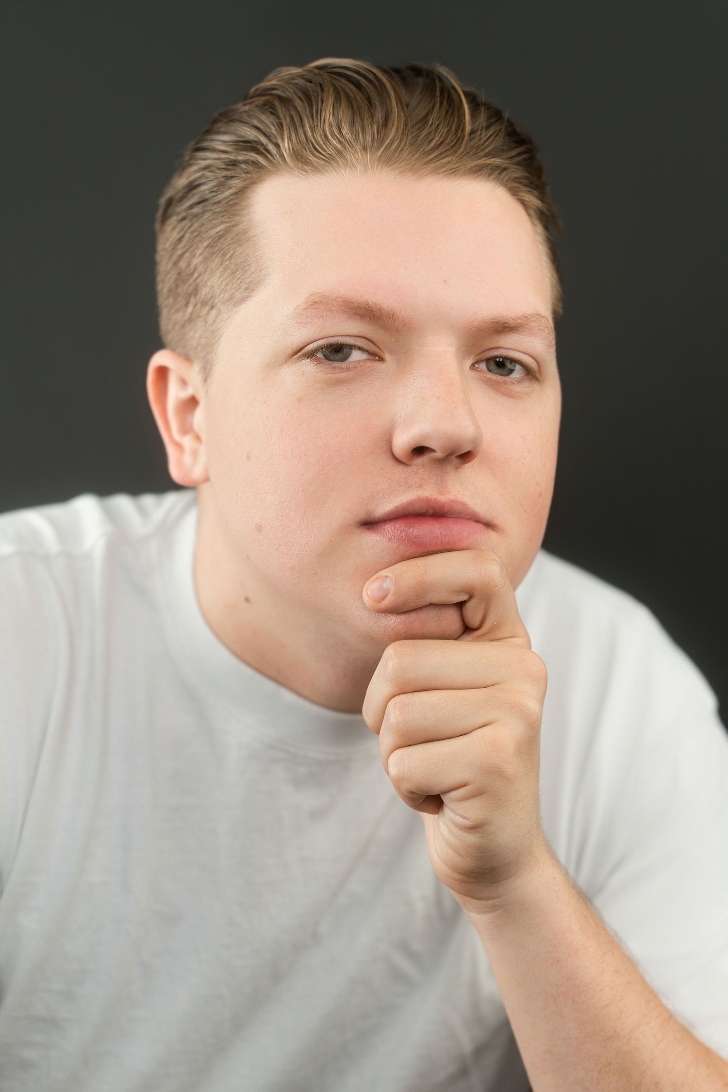 Young man in white t-shirt resting chin on fist in thoughtful pose against dark background.