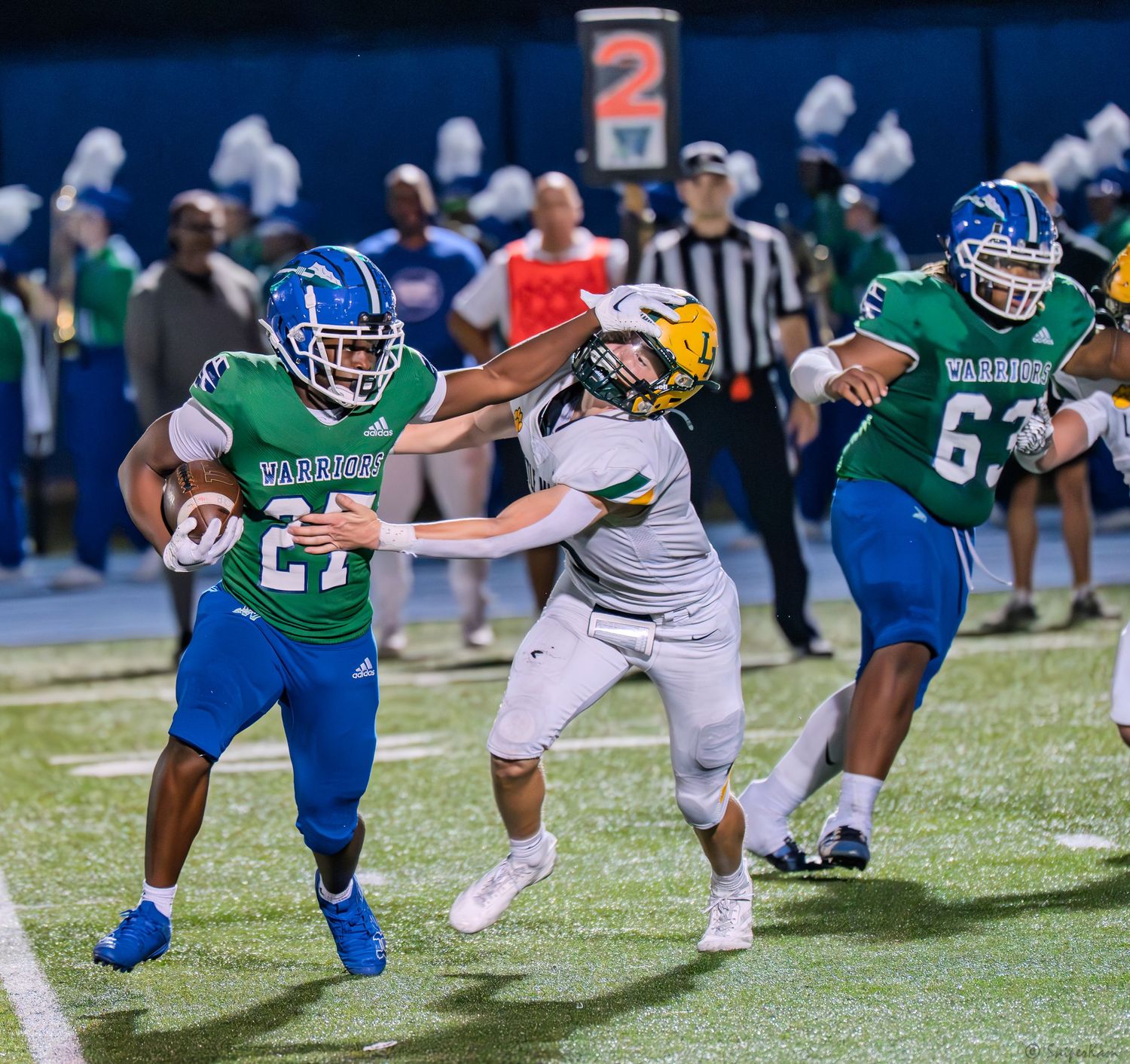 A football player in green and blue uniform runs with the ball while evading defenders during a high school game.