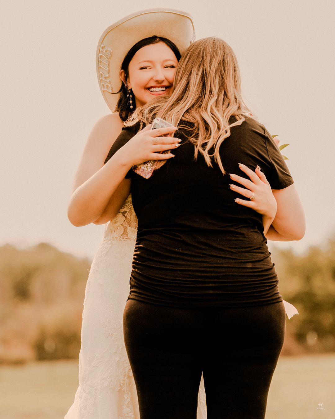Bride in white lace dress and cowboy hat embracing guest in black outfit at outdoor wedding.