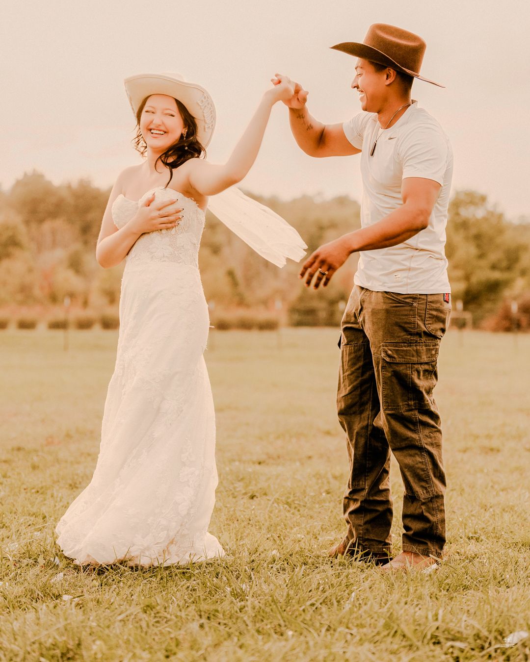 Bride in white dress and cowboy hat being twirled by groom in golden outdoor field.