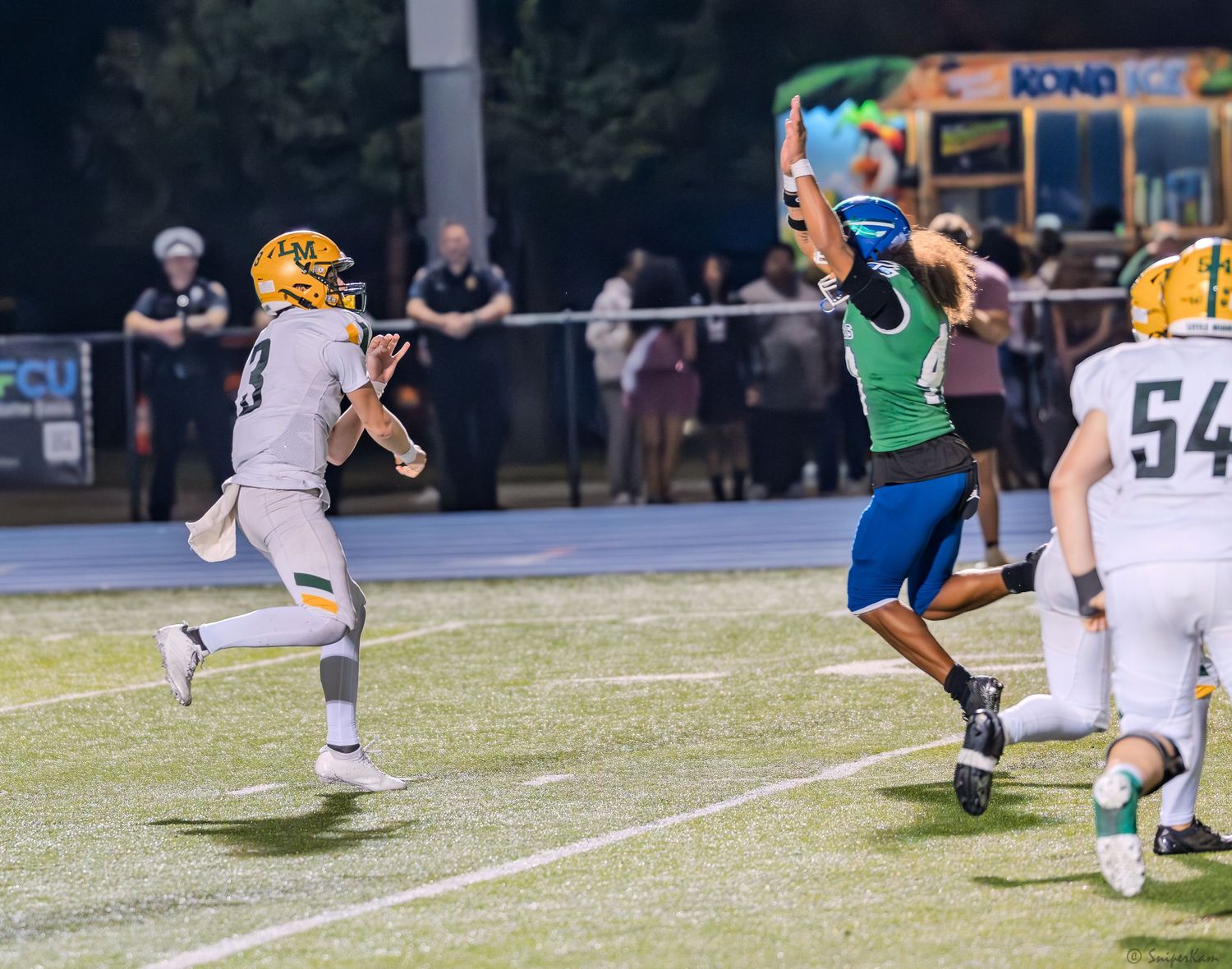 High school football player in green uniform leaps to defend pass during nighttime game.