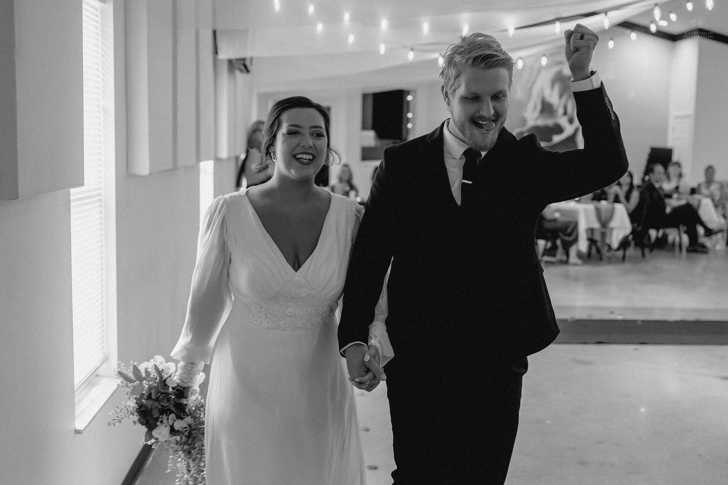Joyful newlyweds walk hand in hand as groom raises fist in celebration at reception.