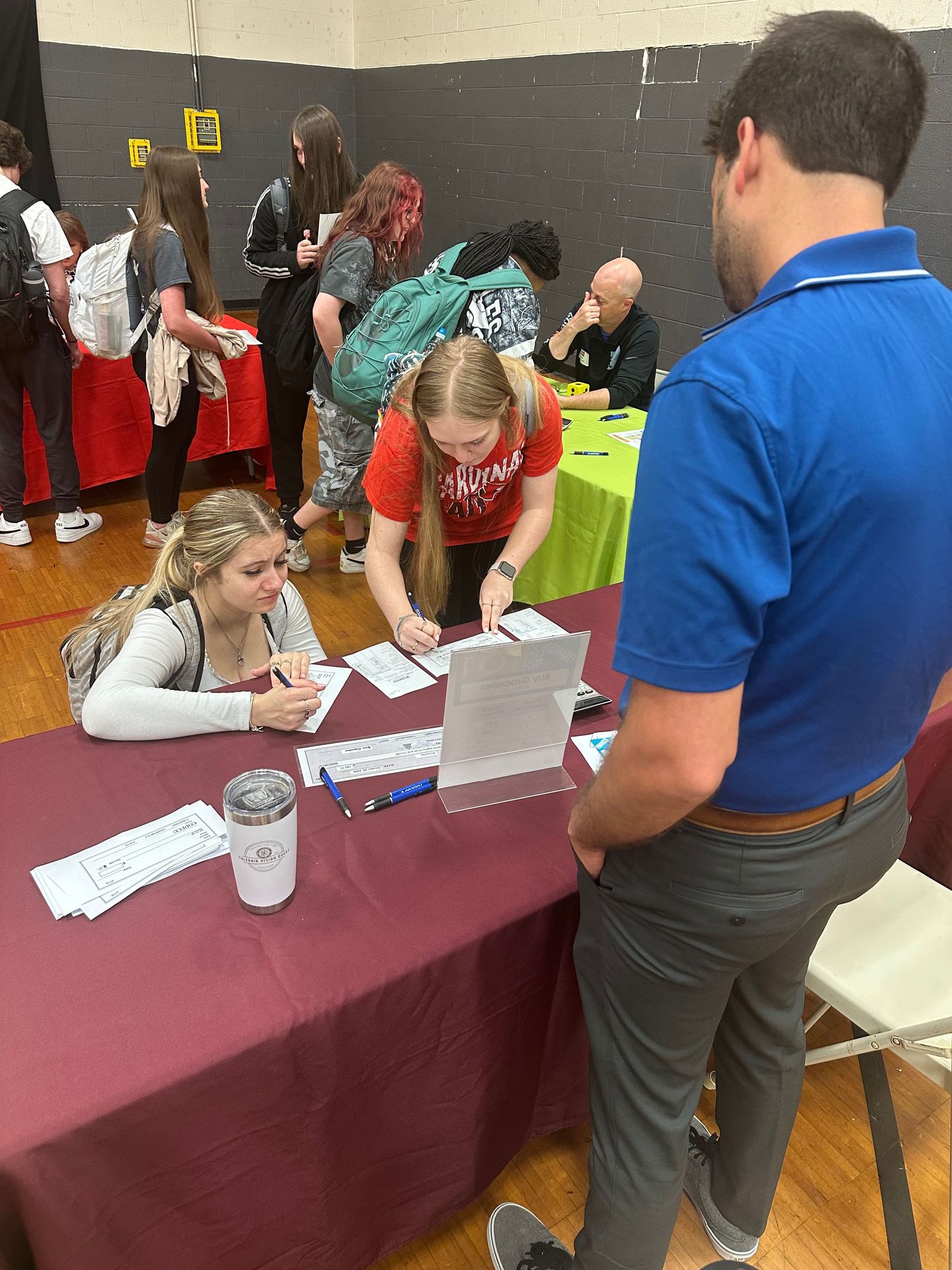 Two female students sign paperwork at a maroon-draped table while a man in blue polo observes.