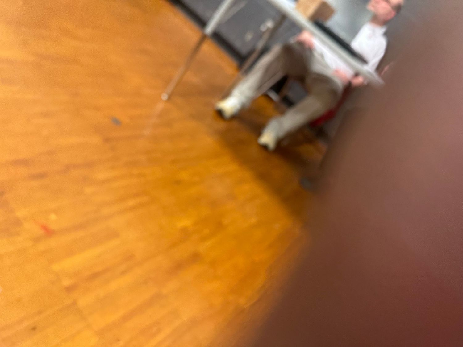 Blurry close-up of a wooden gymnasium floor with a person seated in a chair in the background.