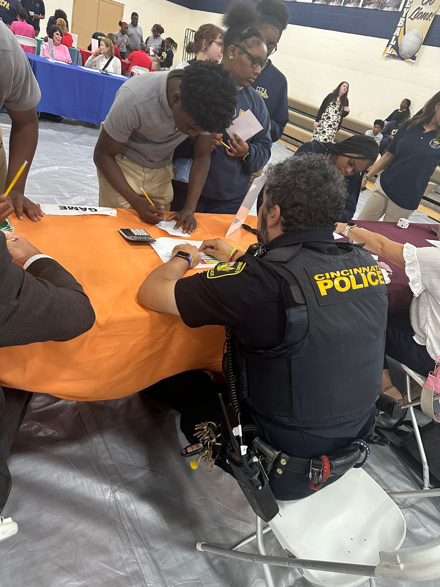 Cincinnati Police officer engages with students at an indoor community event, sitting at an orange table.