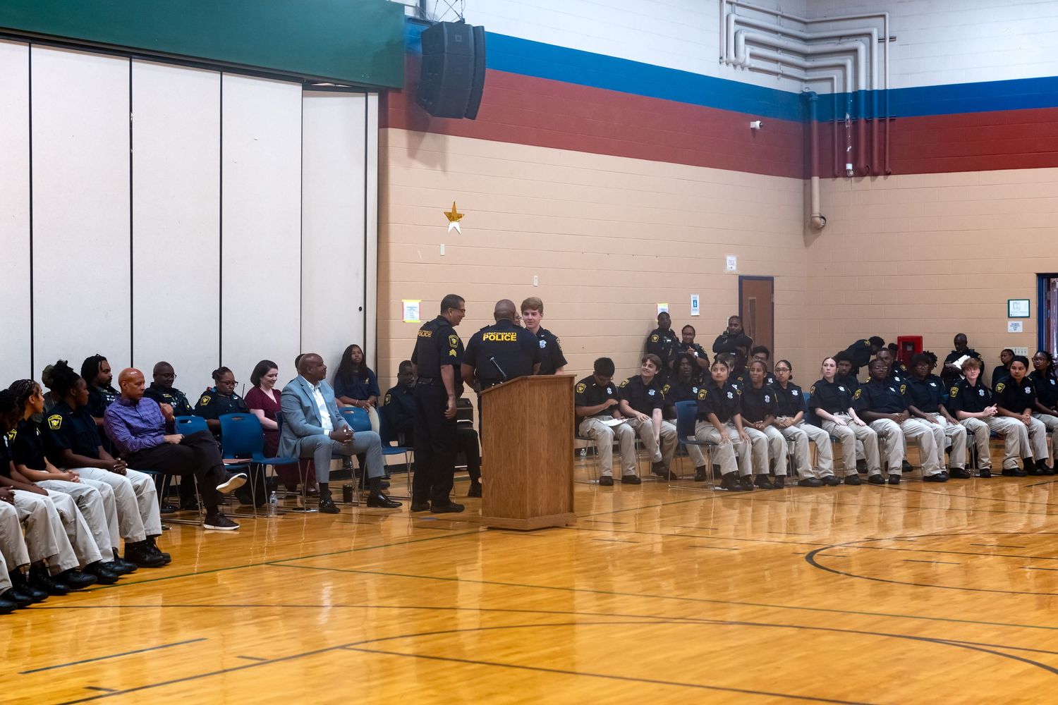Community gathering in a school gymnasium with attendees seated along walls while a speaker presents at a wooden podium.
