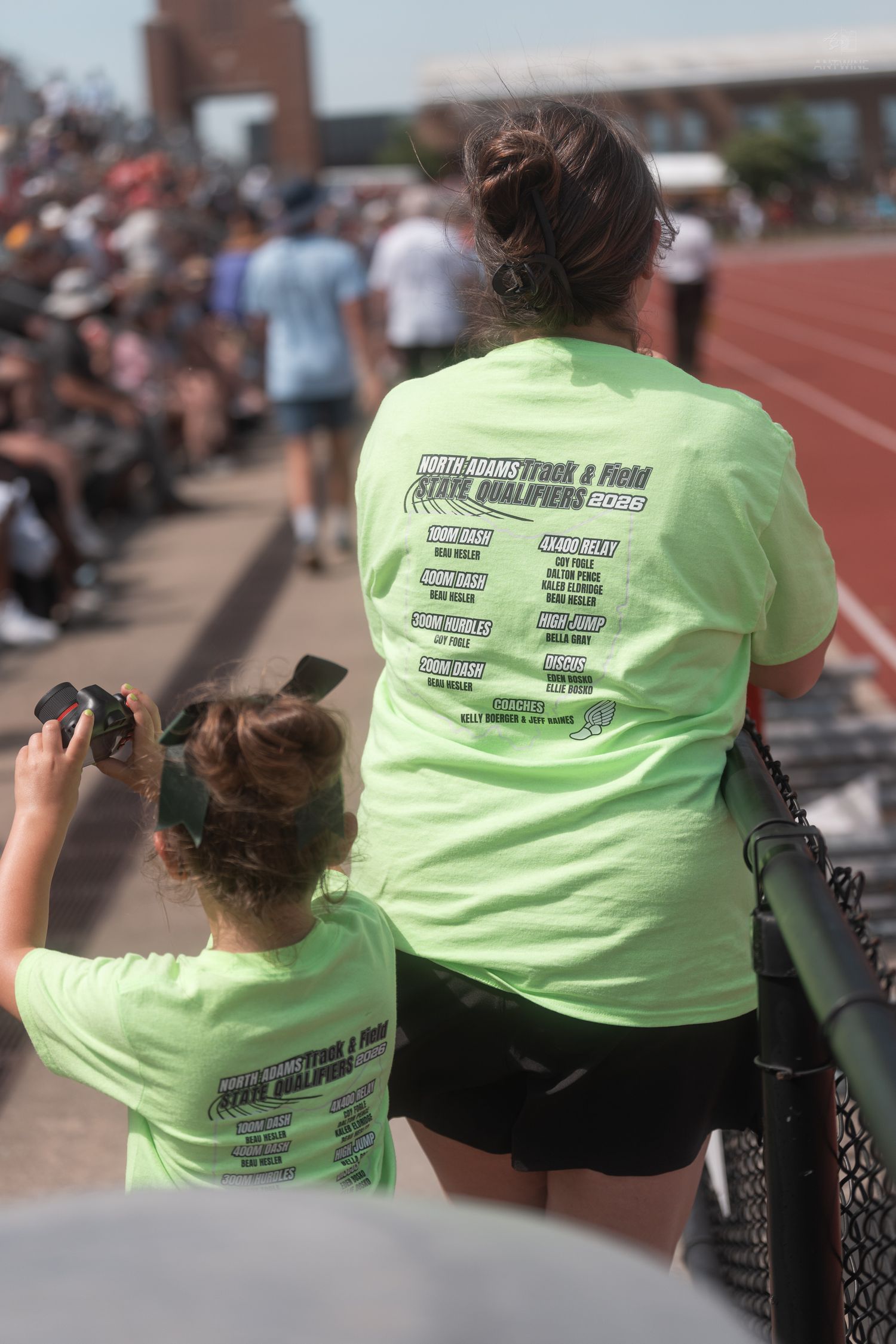Mother and child in matching neon green North Adams Track and Field State Qualifiers shirts watch a race.