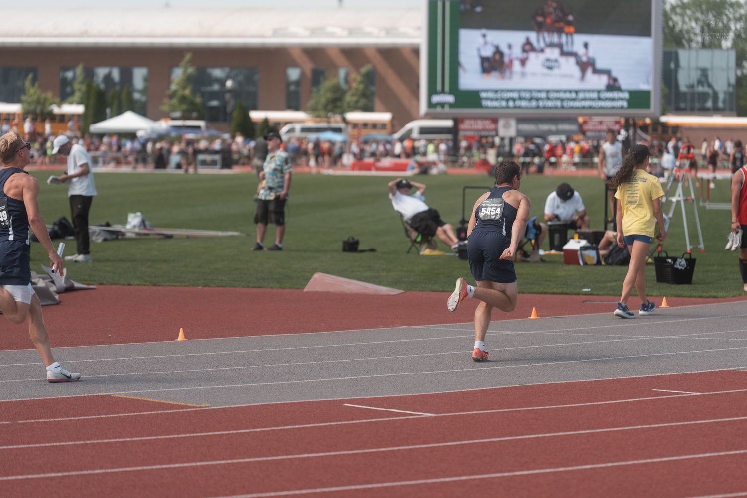 Young athletes compete in a sprint race on a red track at an outdoor high school track and field meet.
