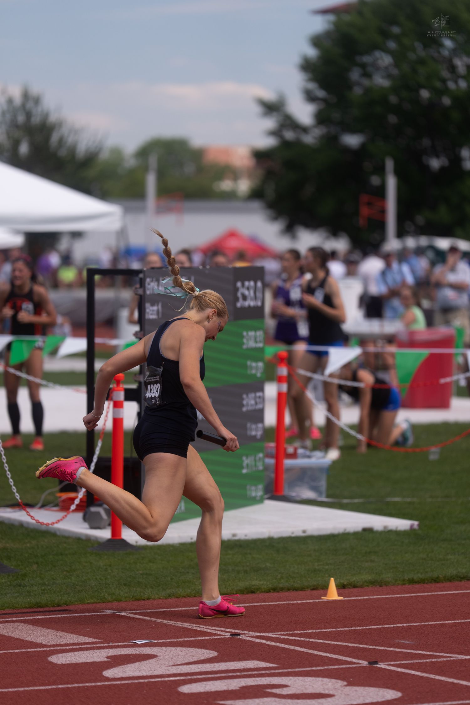 Female athlete in black uniform finishes relay race leg on red track at state championship meet.