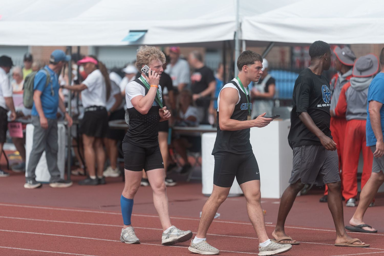Two male track athletes in black and green uniforms walk on a red track at a crowded outdoor meet.