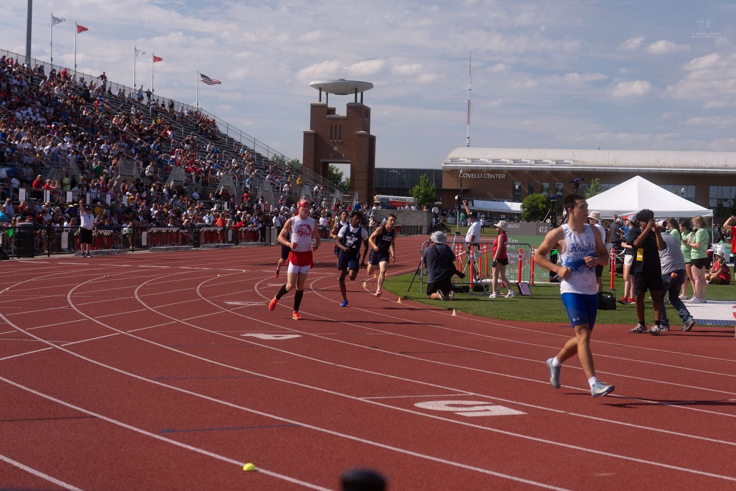 Relay runners exchange batons on a red track during an outdoor high school track and field championship event.