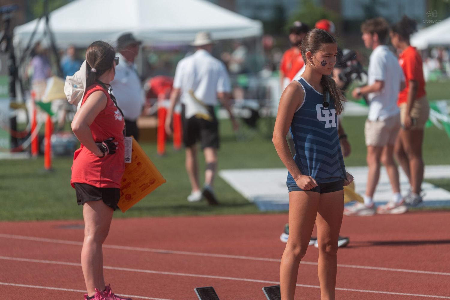 Two female athletes in GH uniforms stand on a red track during an outdoor track and field event.