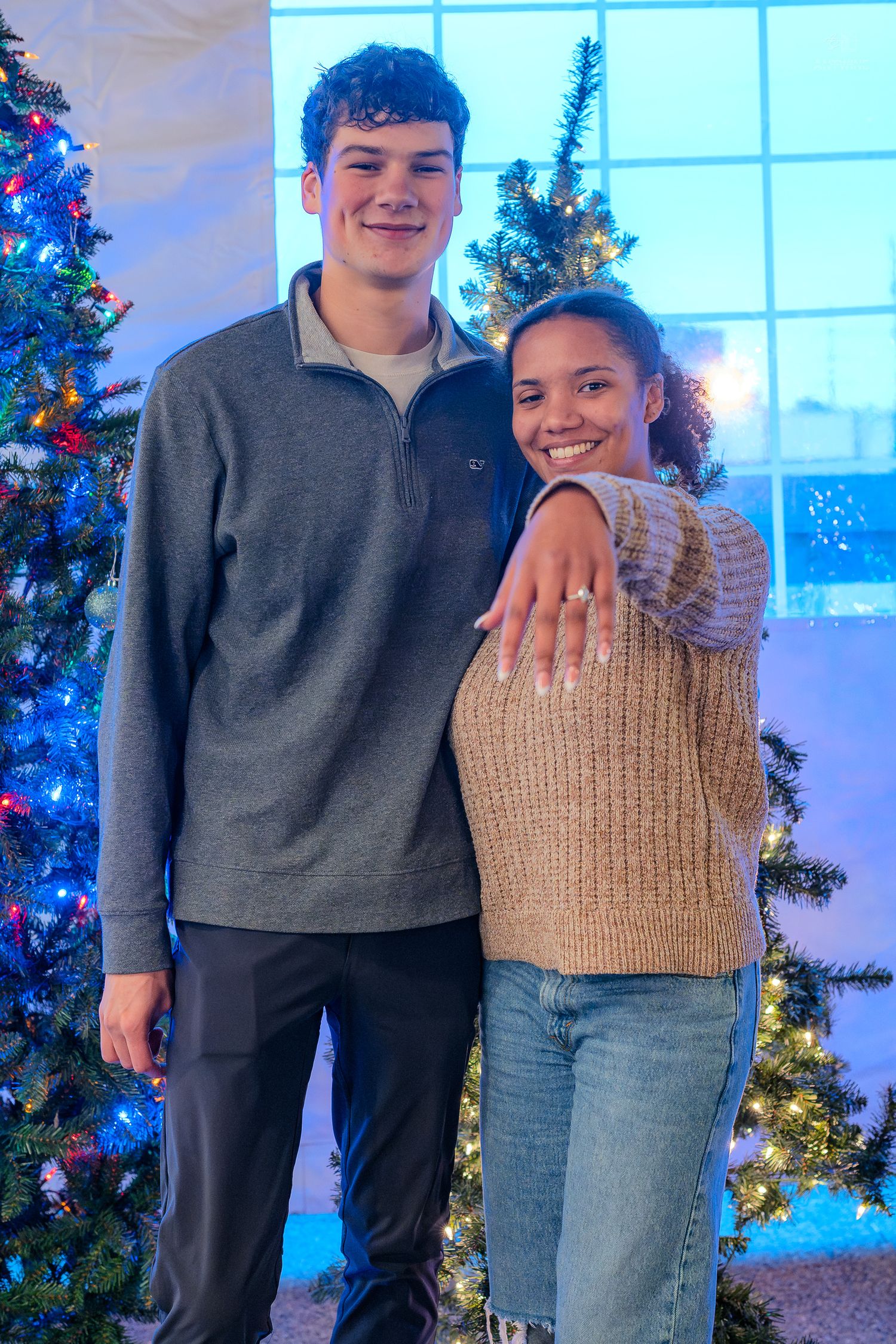 Smiling couple stands together near decorated Christmas tree, woman displaying ring.