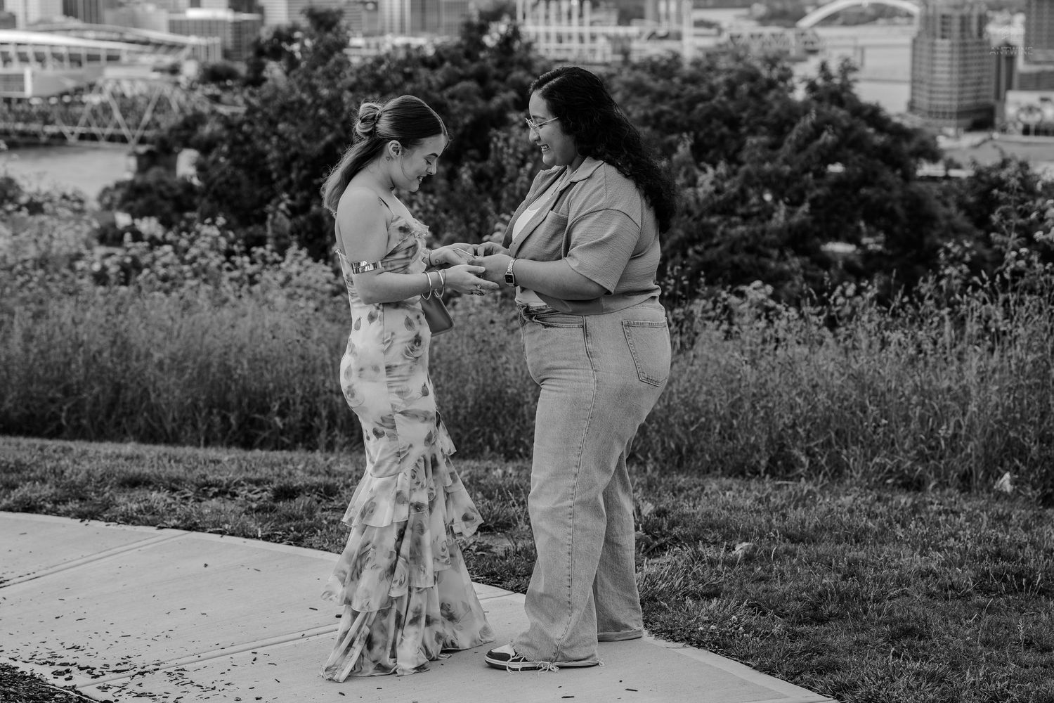 Two women sharing a tender moment holding hands on outdoor pathway with greenery in black and white.