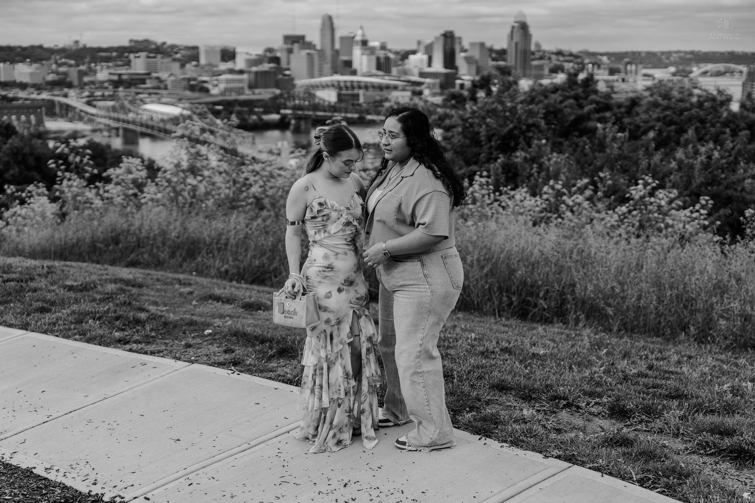 Two women stand close together on a scenic overlook with a city skyline in the background.