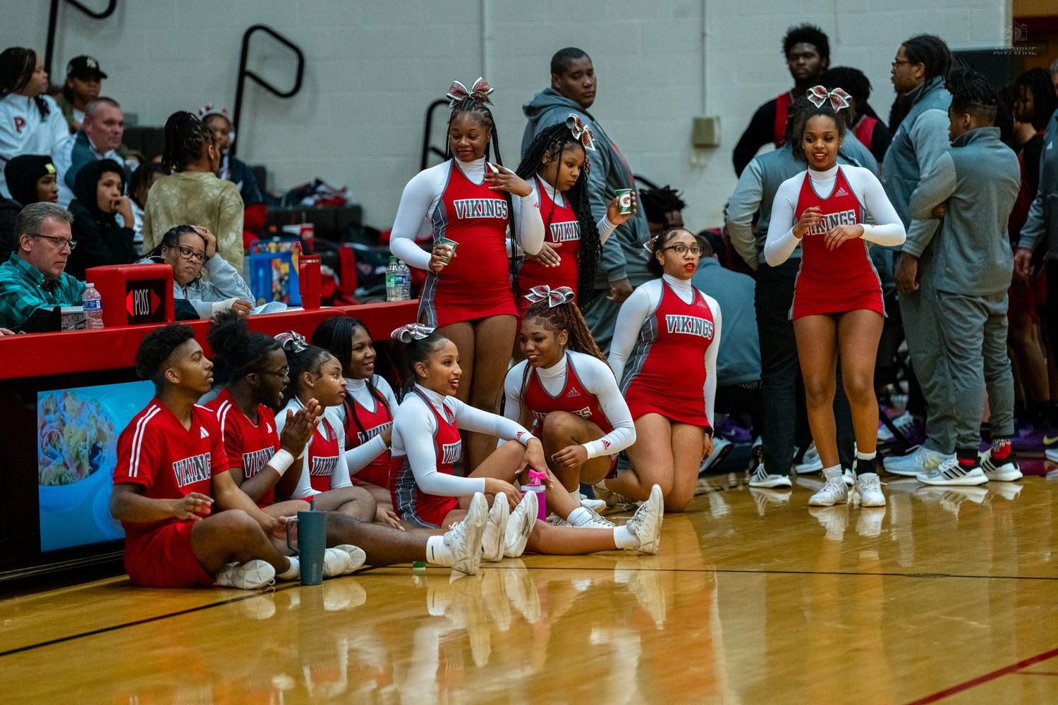 High school cheerleaders in red and white uniforms sit courtside during a basketball game.