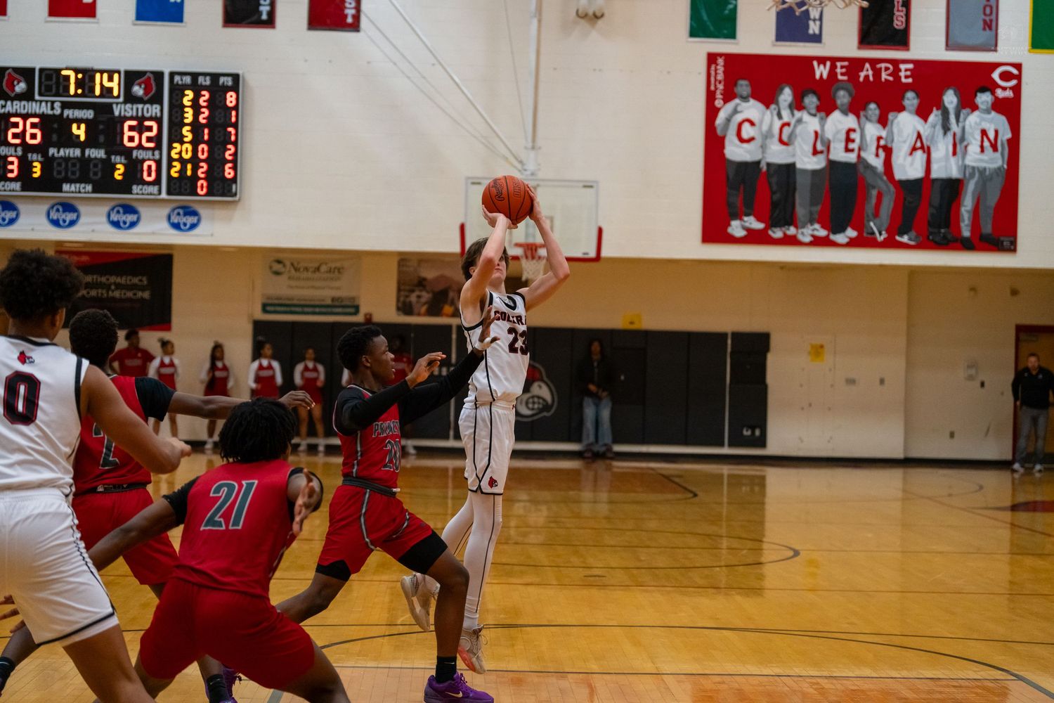 High school basketball player attempts jump shot over defenders during game with scoreboard showing in background.