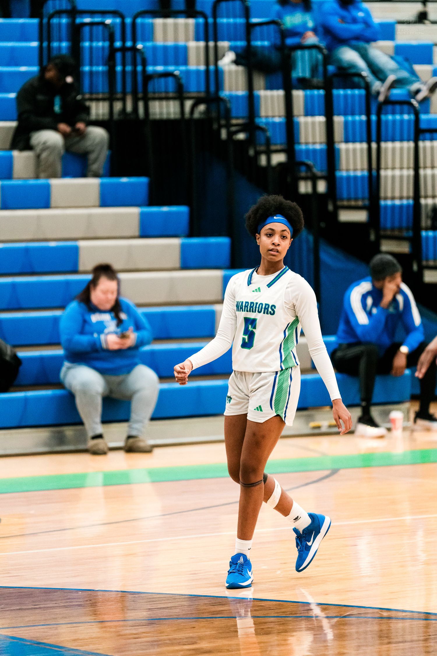 Female Warriors basketball player #5 walks on court in white and teal uniform during high school game.