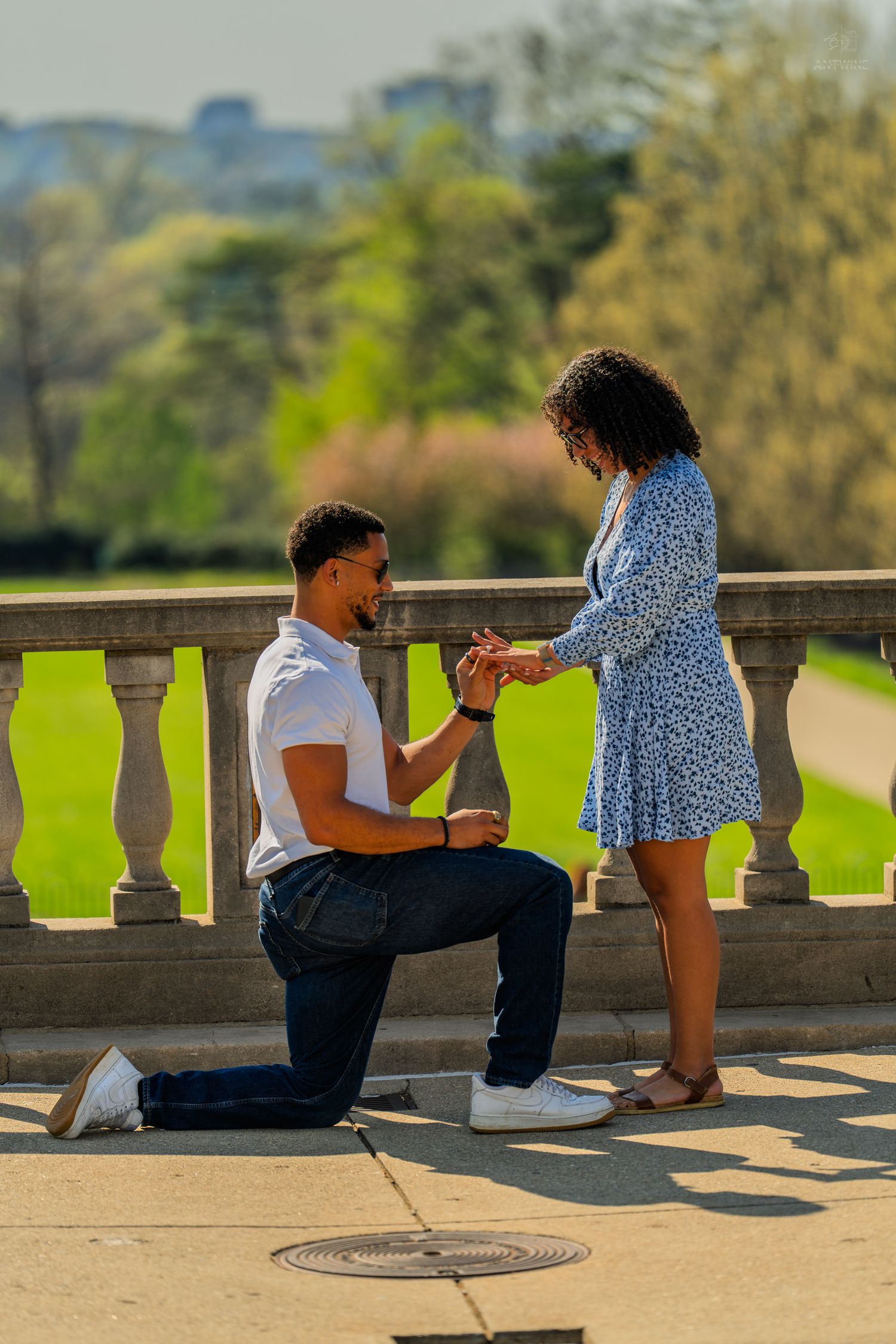 Man kneeling on one knee placing engagement ring on womans finger outdoors in sunny park.